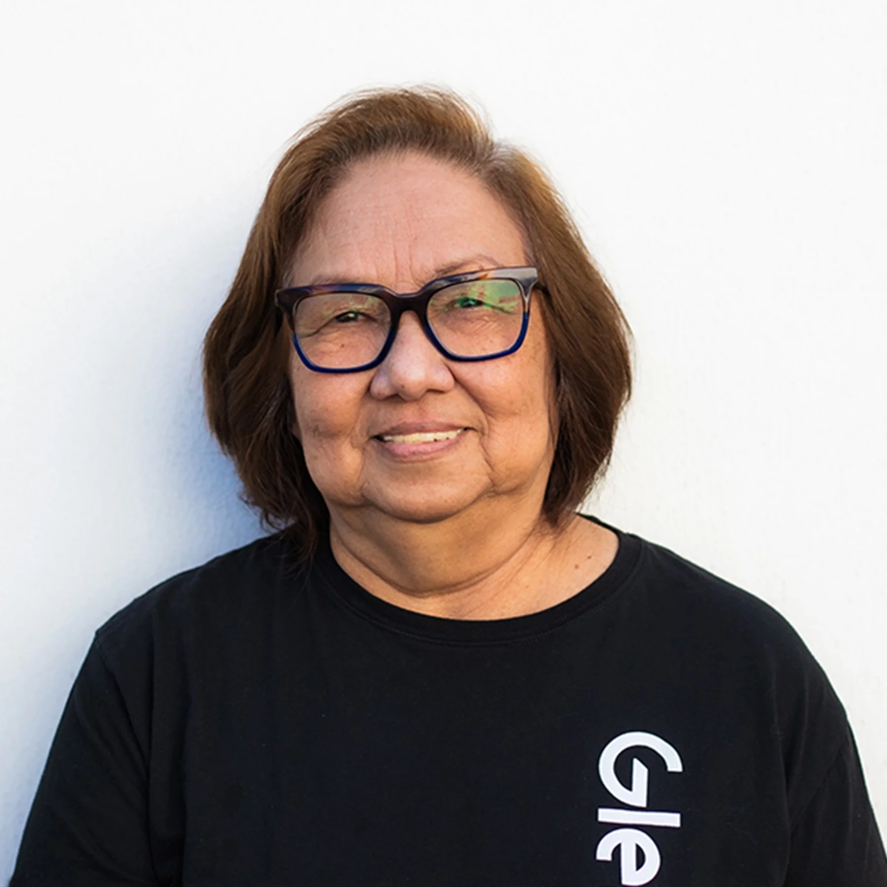 Close-up photo of a smiling woman with reddish-brown hair and glasses, wearing a black T-shirt with white text, standing against a plain white background.