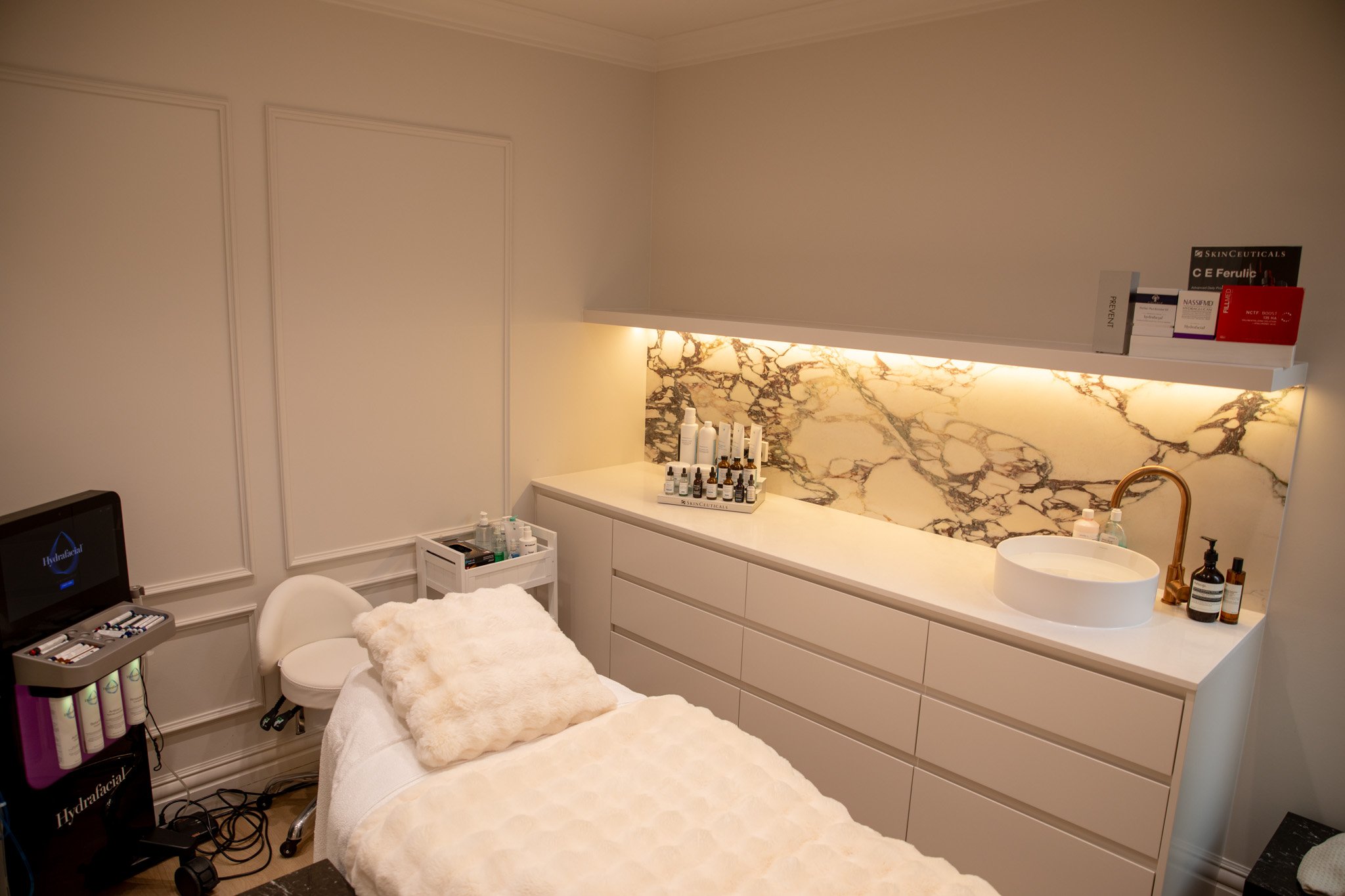 A skincare clinic treatment room with a massage or facial bed covered in a white towel and soft blanket, white cabinetry with skincare products, a marble backsplash, a sink with a gold faucet, and various skincare products and tools on shelves and counters.