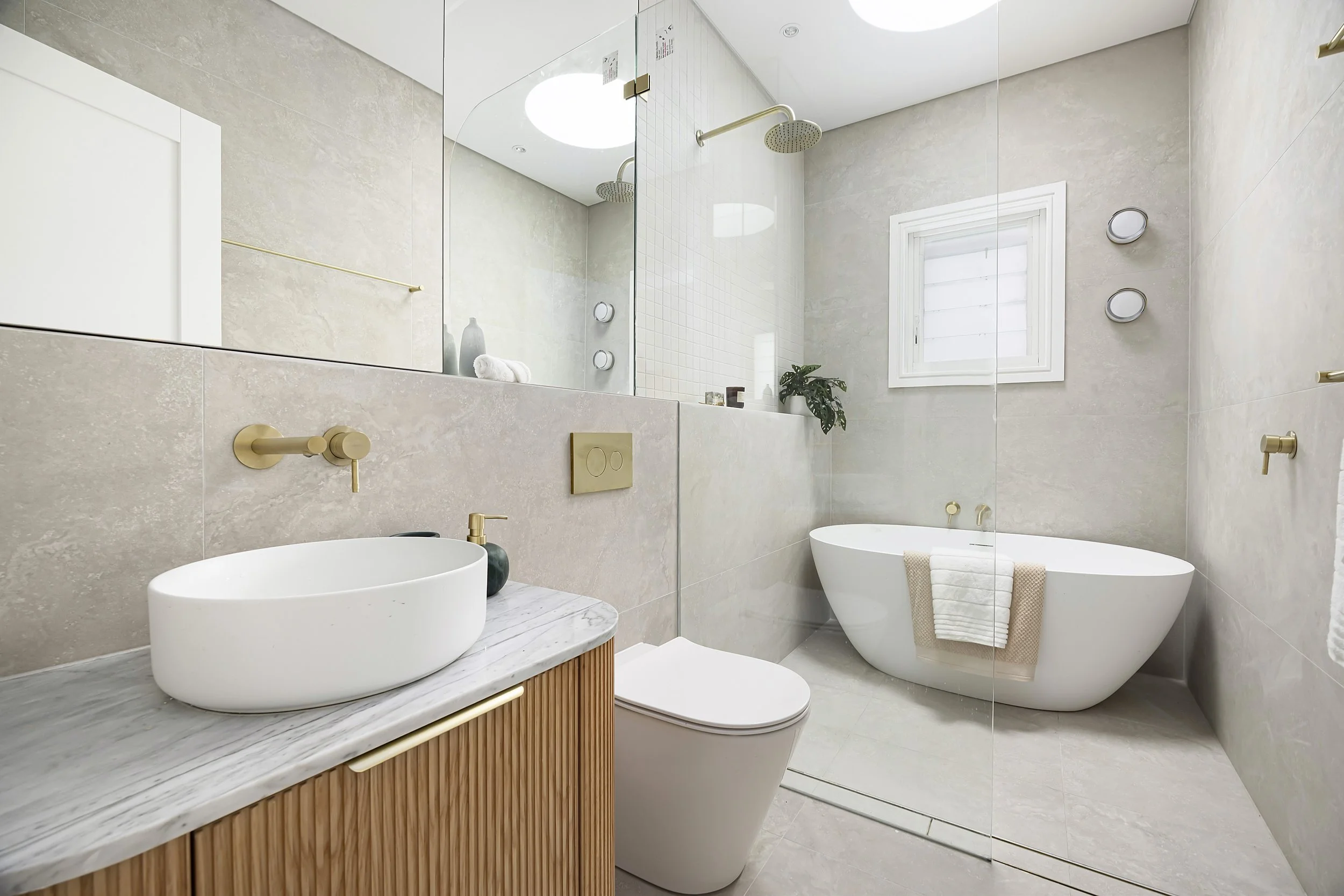 A modern bathroom with a marble top vanity, vessel sink, wall-mounted gold faucet, a toilet, a bathtub, a large mirror, a window, potted plant, and gold fixtures.