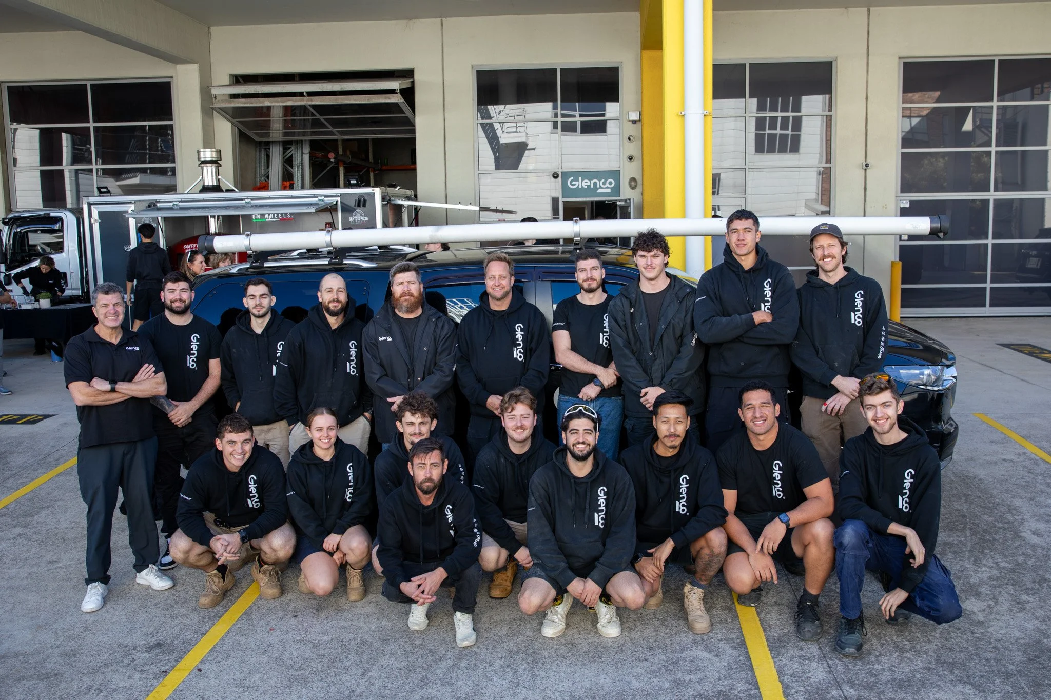 Group of people, Glenco Electrical team posing outdoors in front of a facility with large windows and pipes, some standing, some kneeling, smiling at the camera.