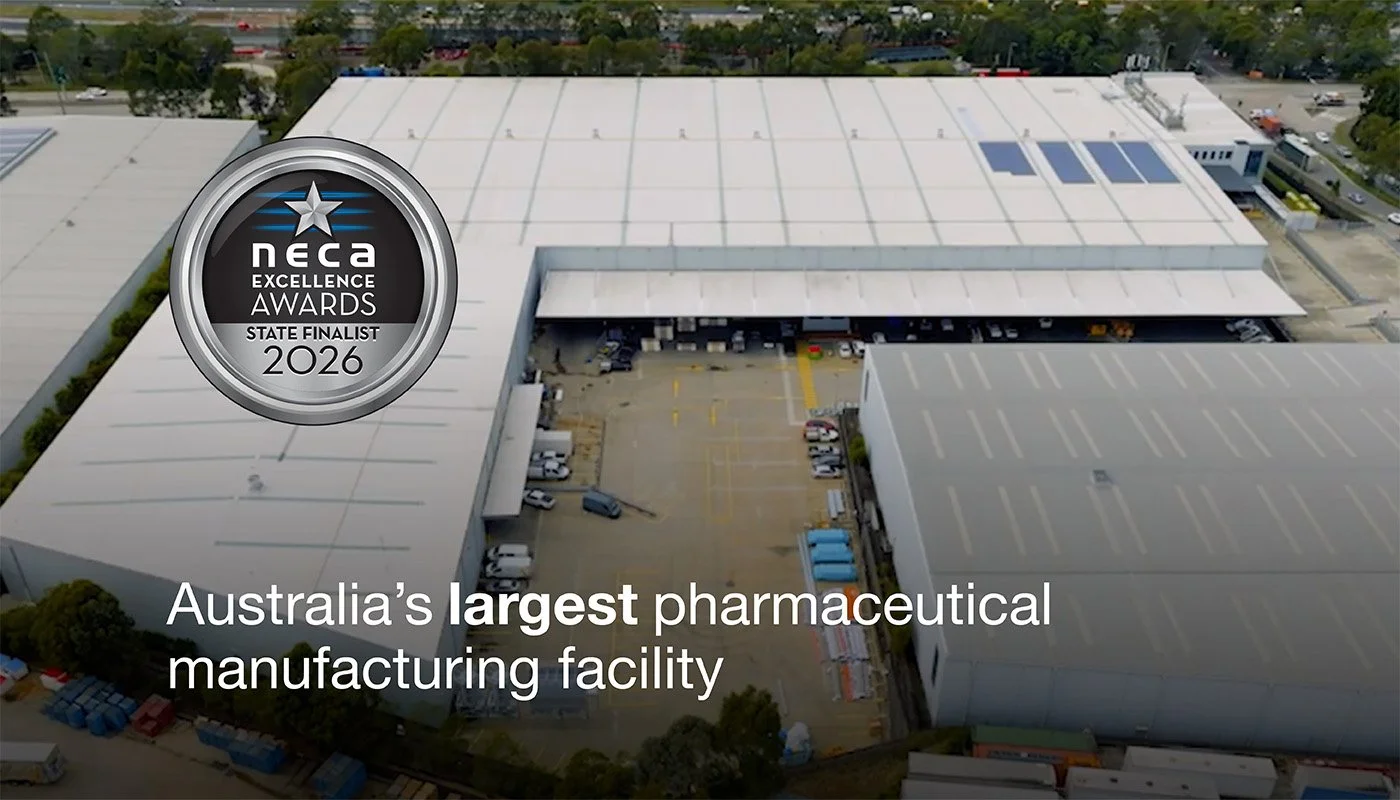 Aerial view of Australia's largest pharmaceutical manufacturing facility, showing large white warehouse buildings, parking lots, and surrounding greenery. Contains a NECA Excellence Awards 2026 state finalist badge.