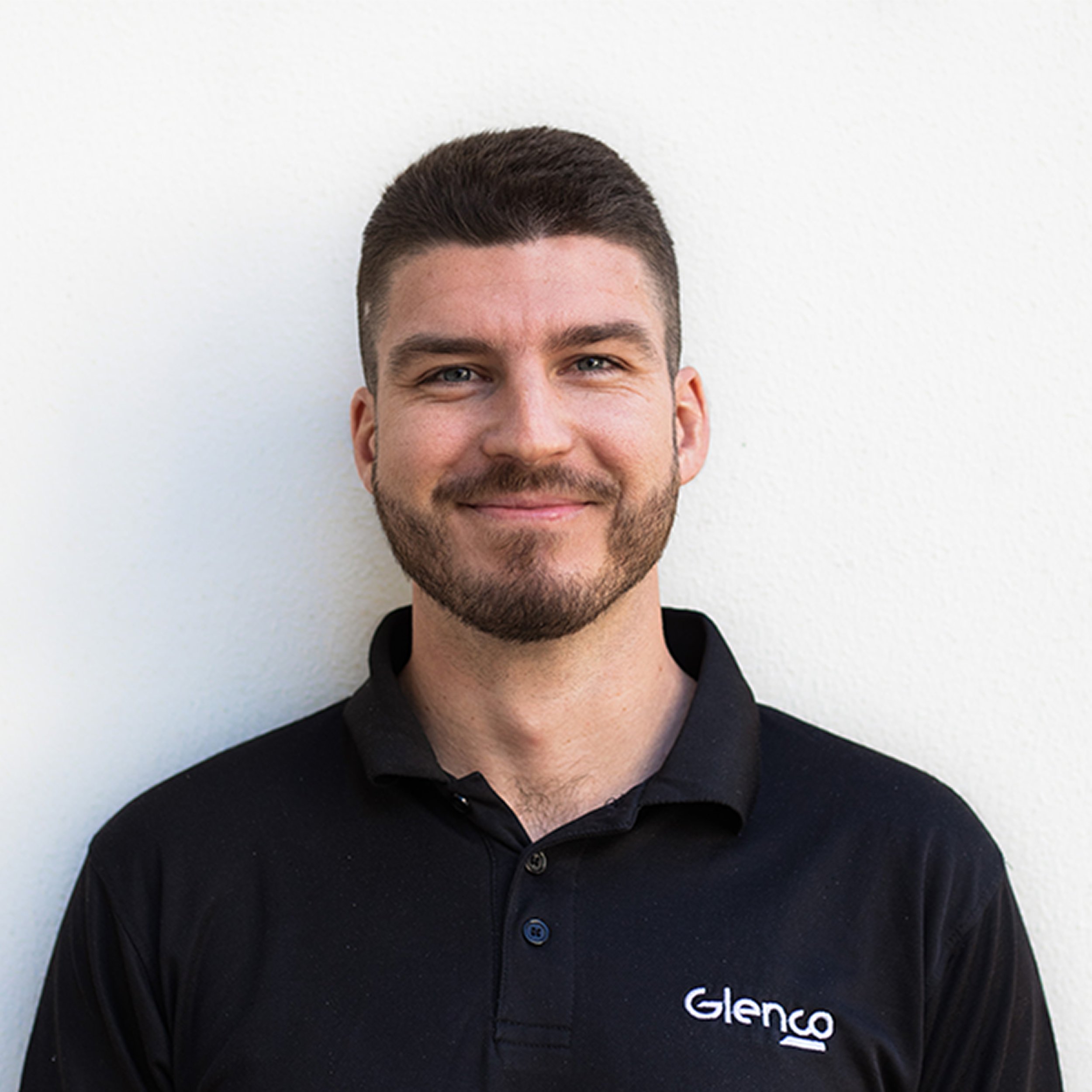 Close-up of a man with brown hair and a beard wearing a black polo shirt with the logo 'Glenco' on it, standing against a plain white background.