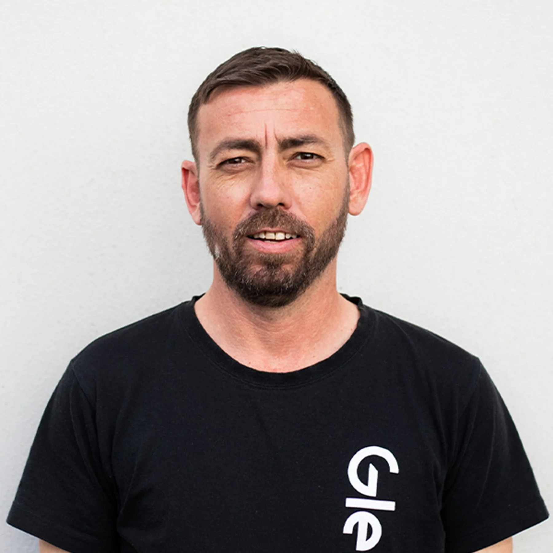 A man with short dark hair and a beard, wearing a black Glenco t-shirt, standing against a plain background
