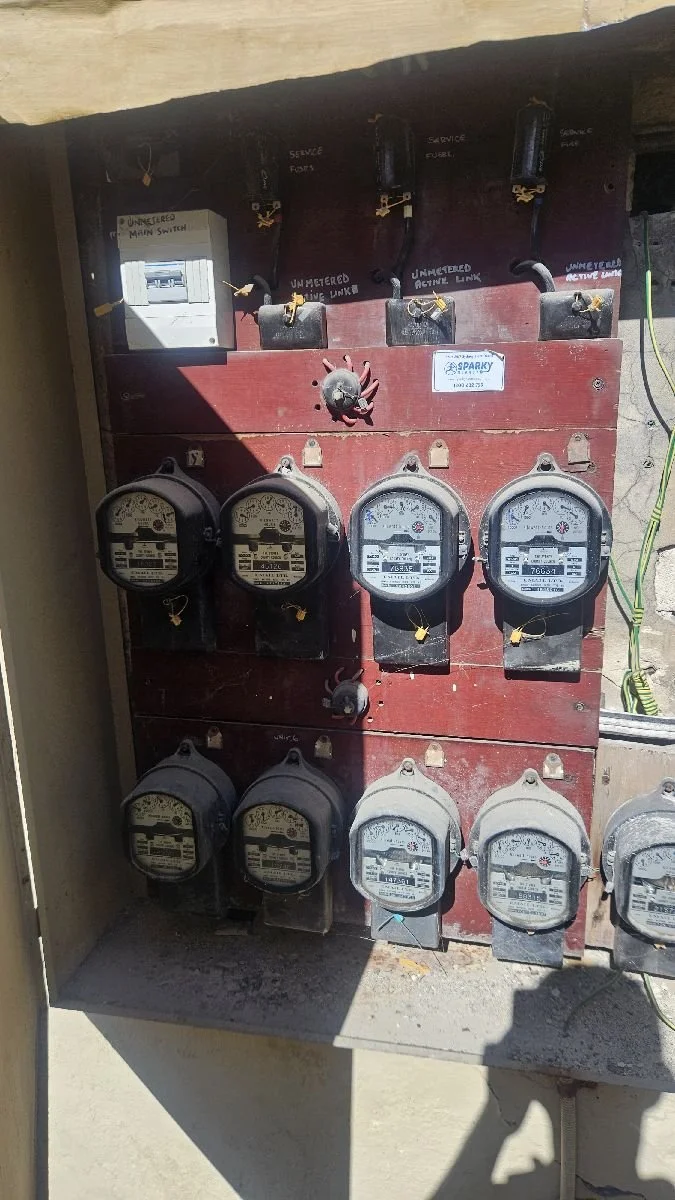 An electric meter panel with multiple meters installed on a wooden board, with some switches and labels above and around the meters.