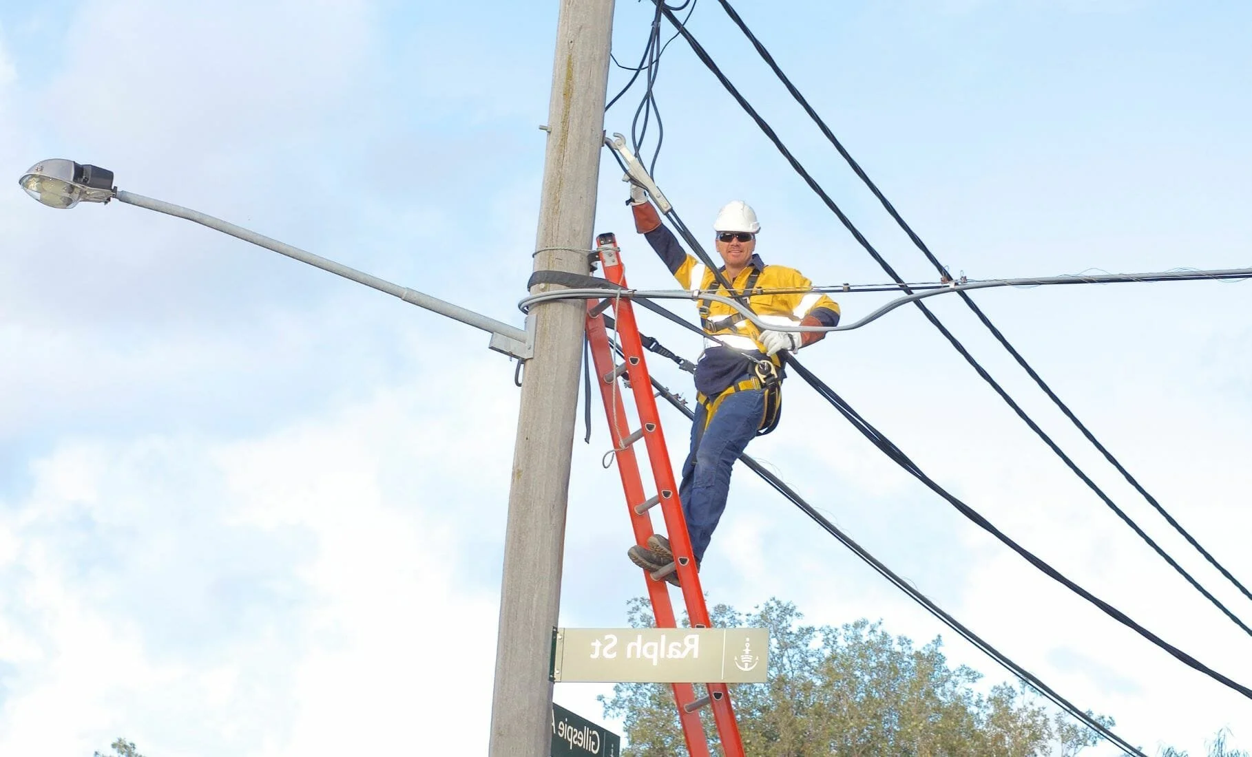 A worker in safety gear, including a helmet and harness, standing on a red ladder, repairing or inspecting power lines on a utility pole during daytime. The worker is smiling and dressed in a yellow and navy uniform with safety gloves and sunglasses.