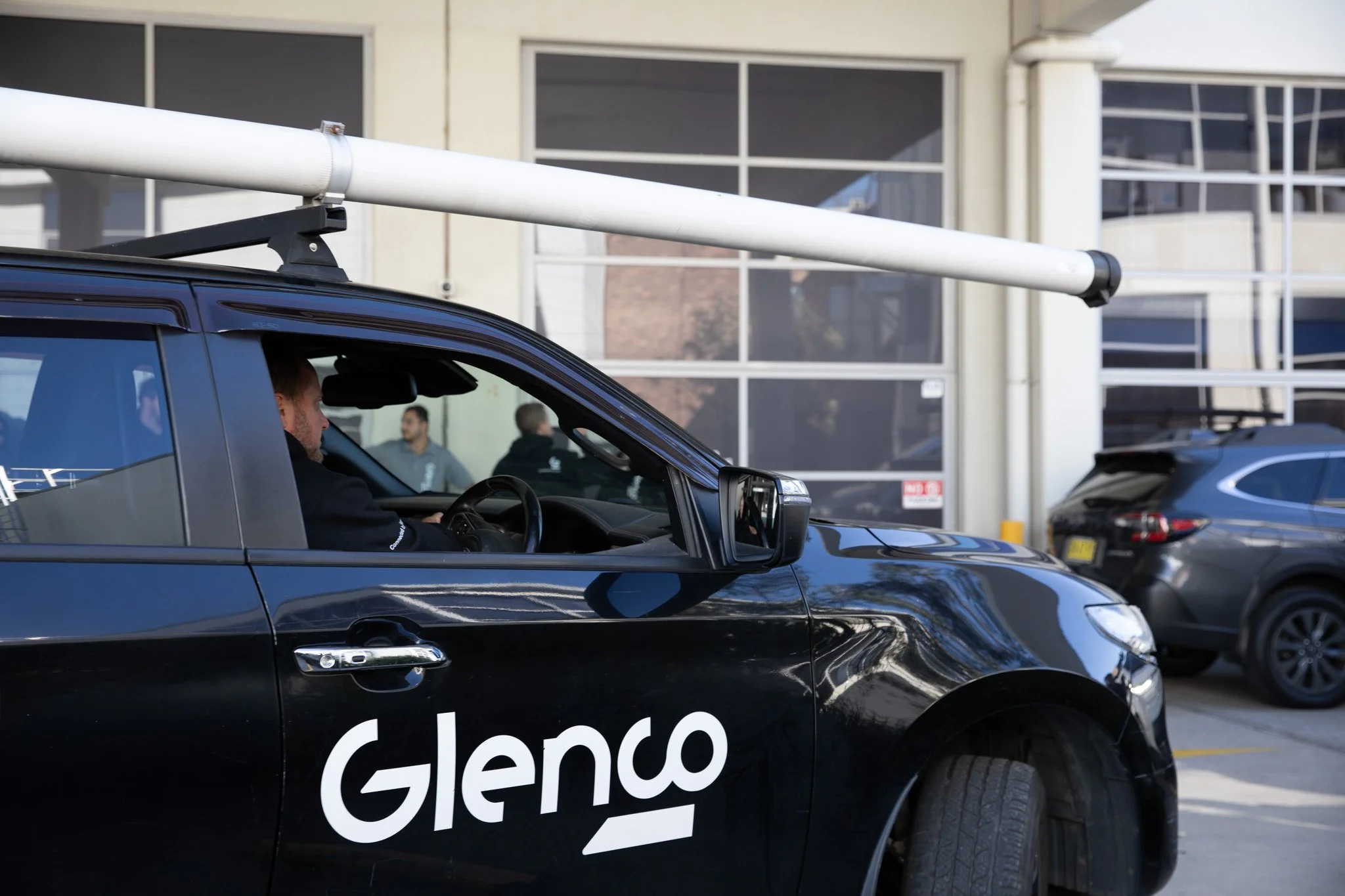 Black vehicle with 'Glenco' logo, roof-mounted equipment, parked in a lot with other cars, in front of a modern building with large glass windows.