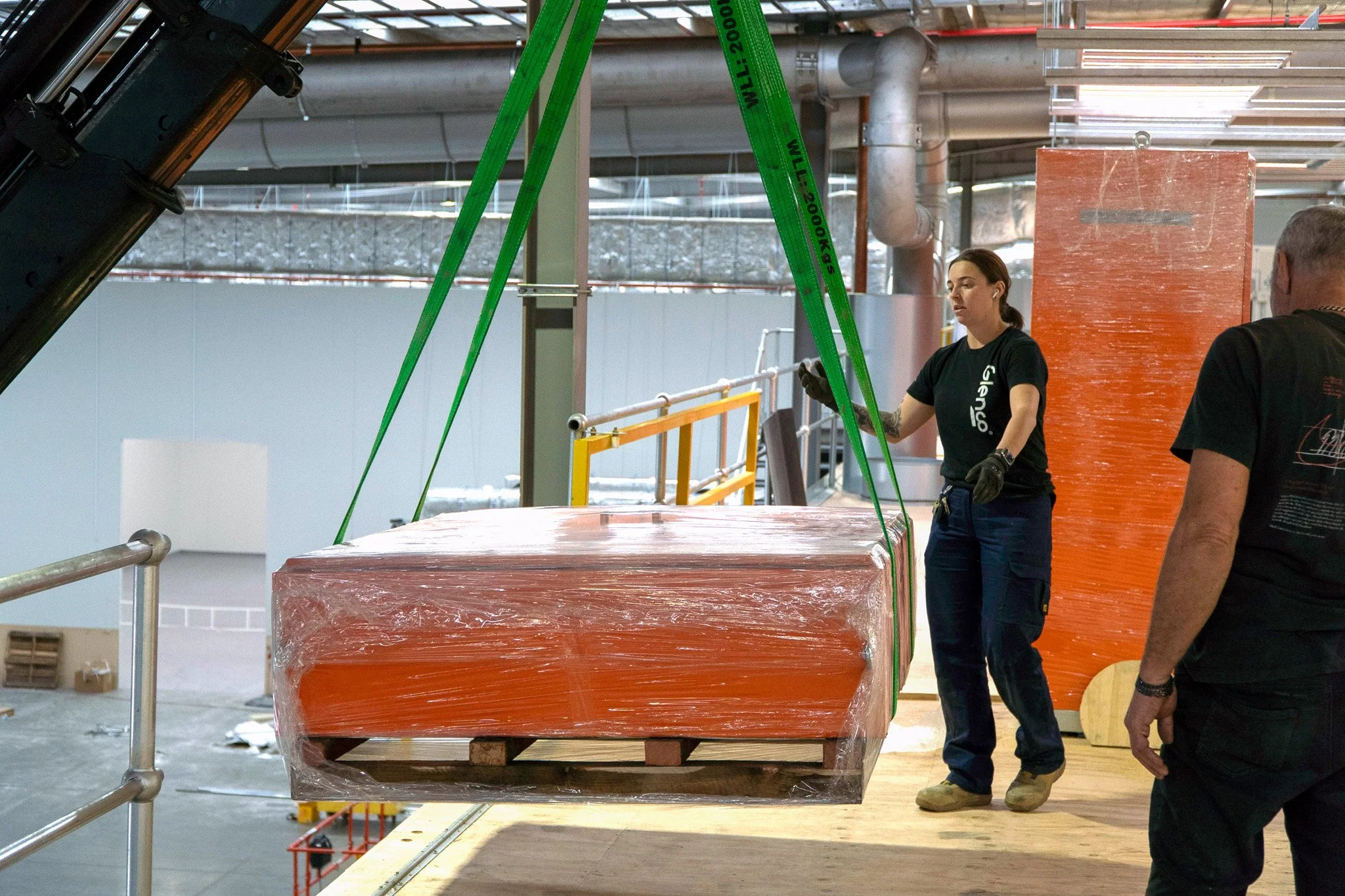 Workers in a factory or construction setting handling large, wrapped orange objects suspended from a crane or hoist system.