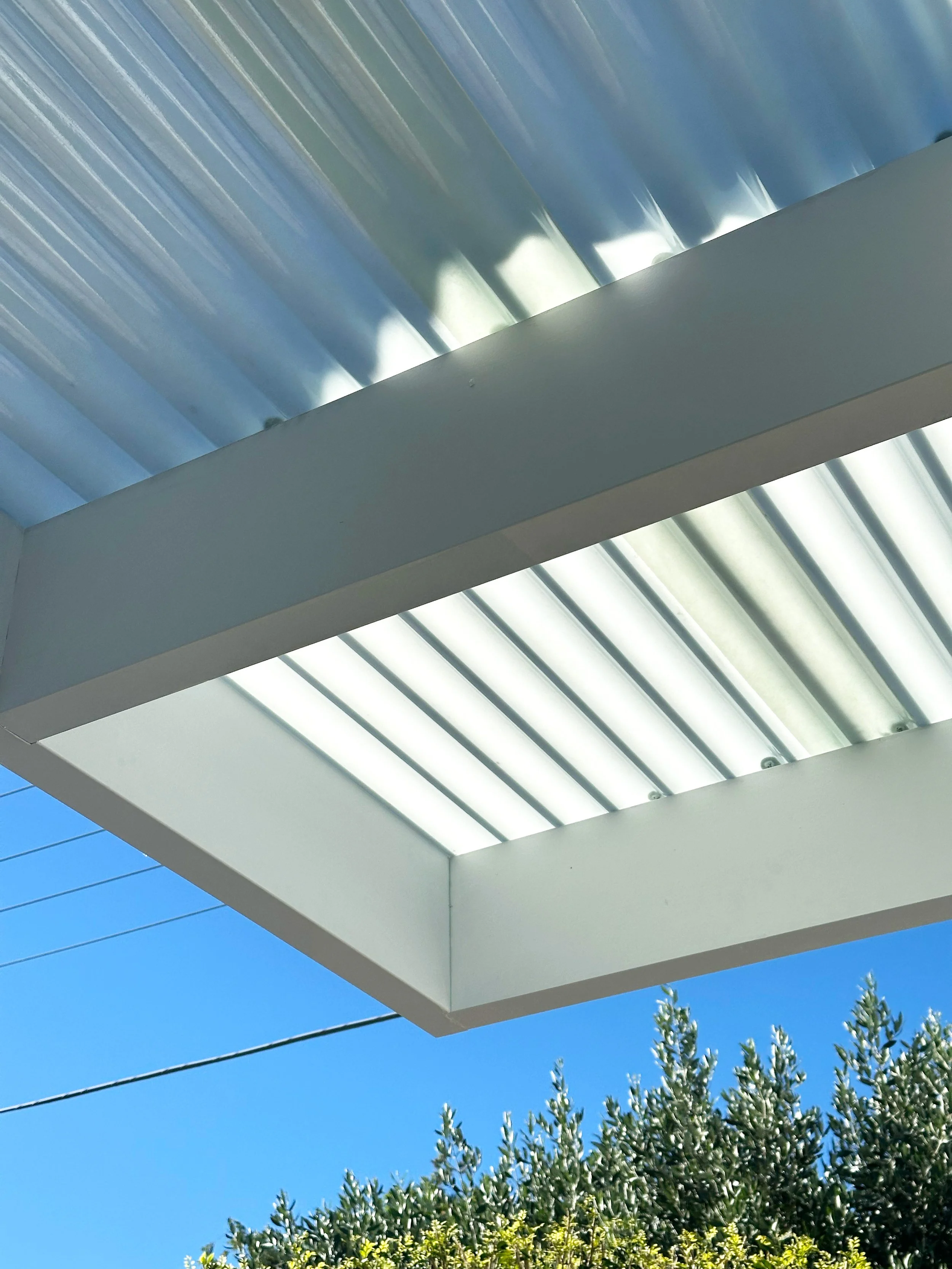 The image shows a modern building with a sunshade or roof, set against a bright blue sky with some tree foliage at the bottom.