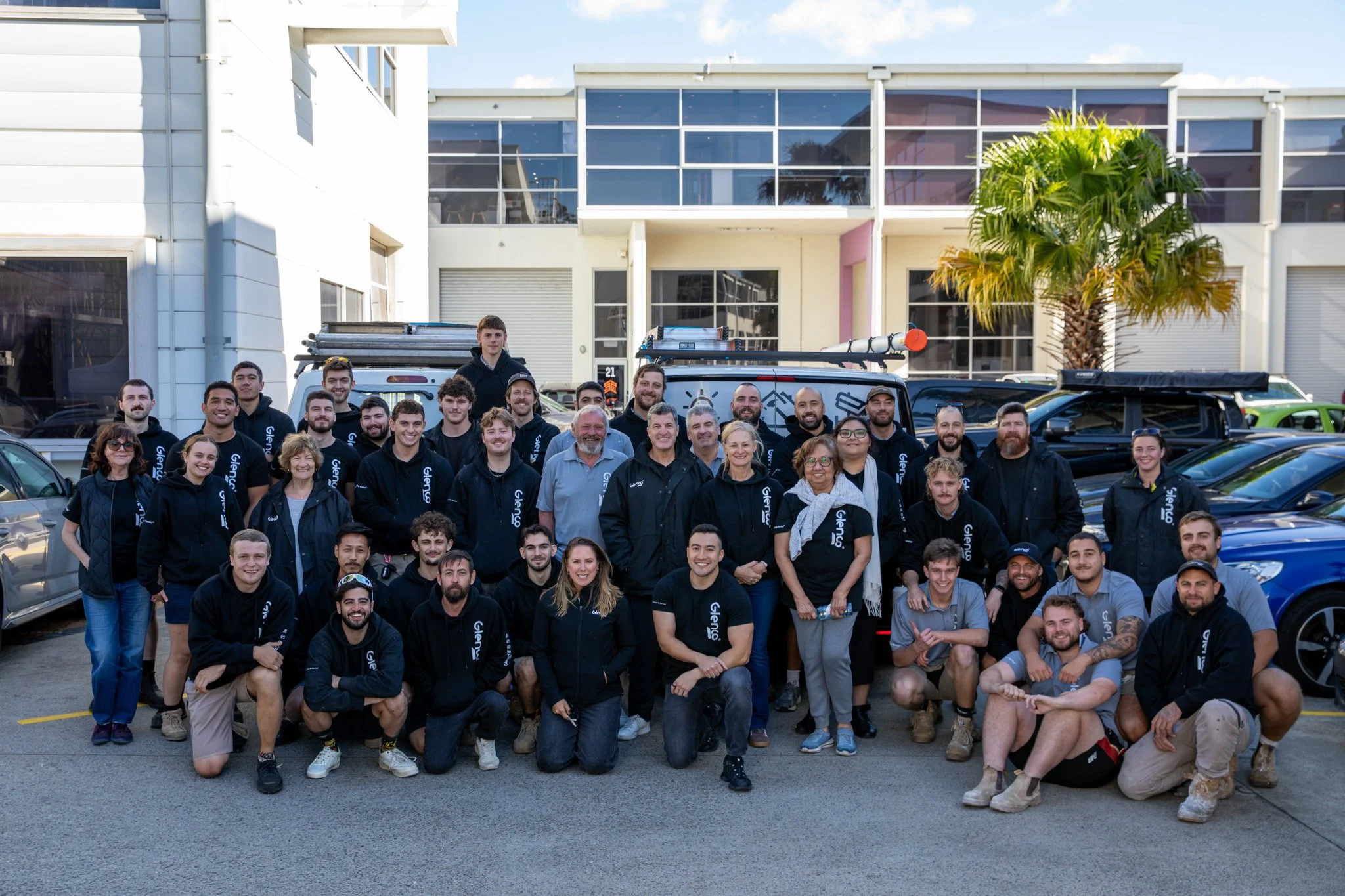 A large group of people standing outdoors in front of a modern building, some wearing black jackets with the Gleno logo, posing for a group photo. The background features parked cars, a palm tree, and a blue sky with some clouds.