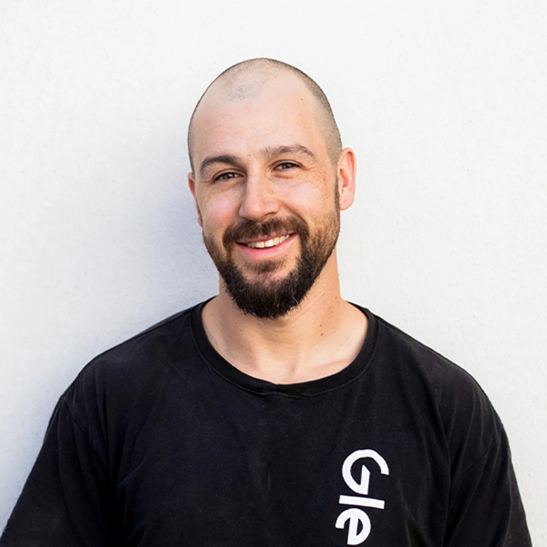 A smiling man with a beard and shaved head standing against a white wall, wearing a black Glenco shirt