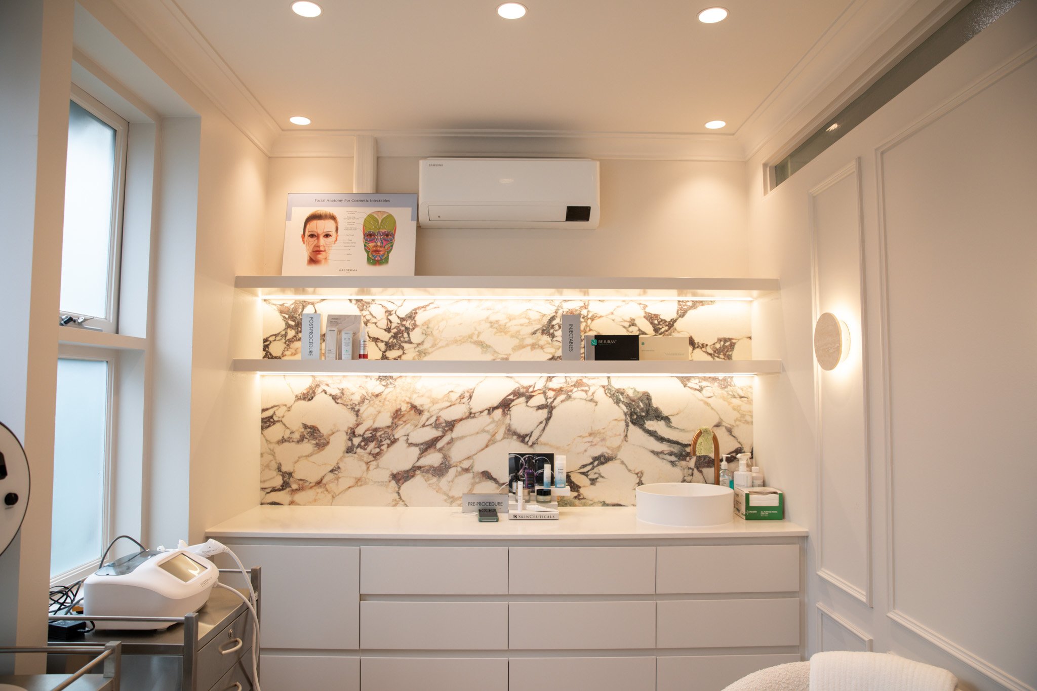 A modern medical or cosmetic treatment room featuring white cabinetry and marble backsplash. On the counter, there are various skincare products and supplies, with a circular sink and a small mirror. On the left, a monitor with medical equipment is visible. The wall above showcases an anatomical facial diagram poster.