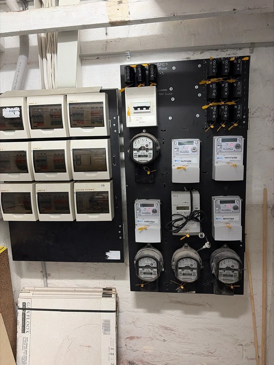 Multiple electric meters and circuit breakers mounted on a black board next to white electrical boxes on a wall.