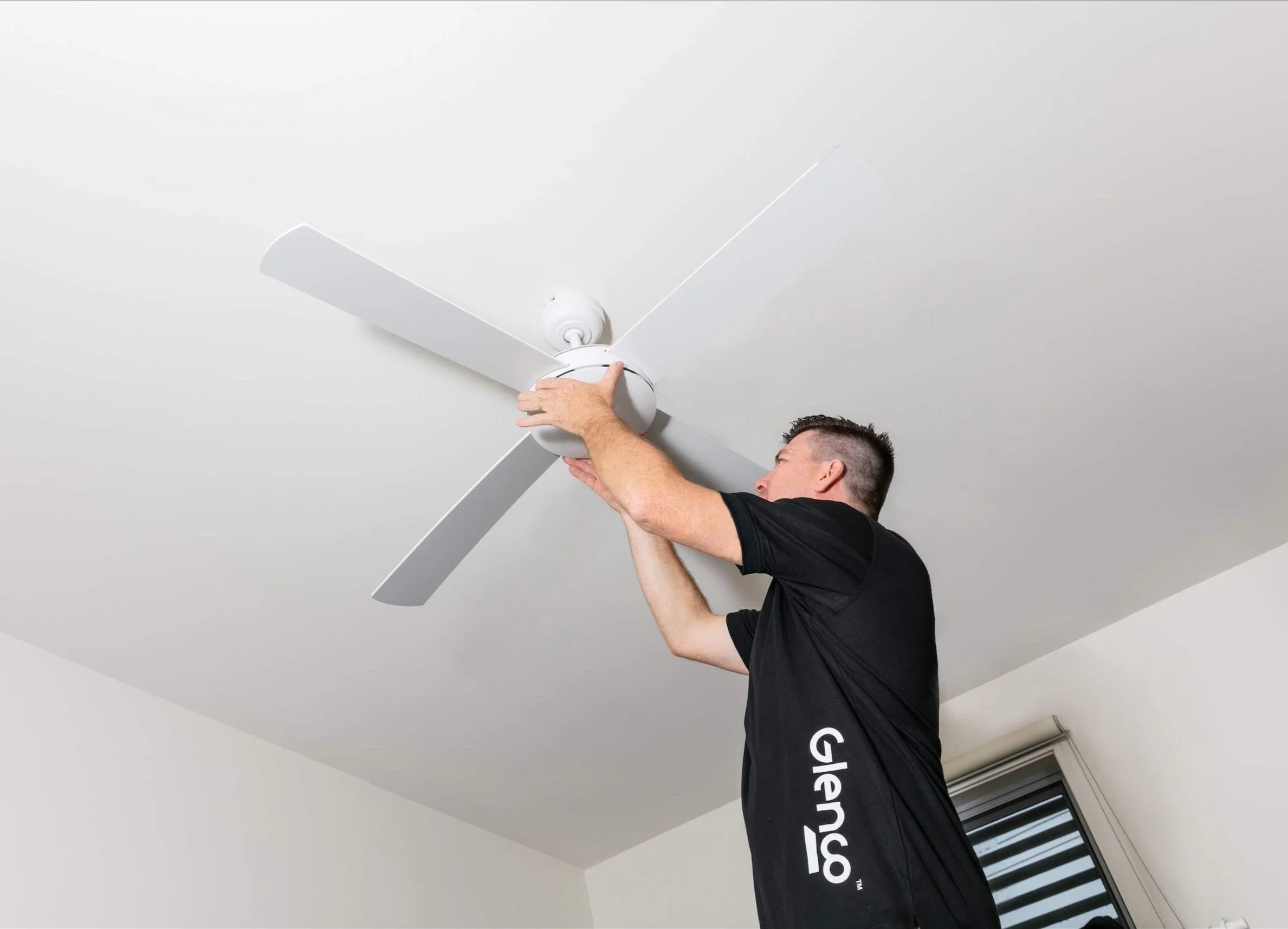 A man wearing a black shirt with white text, installing a white ceiling fan