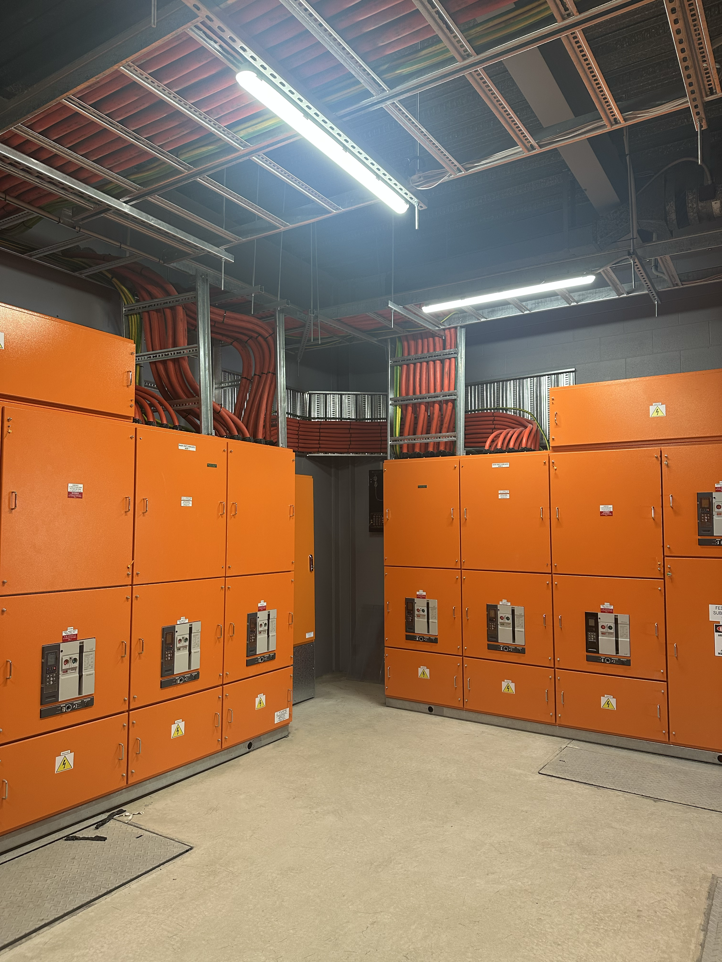 Electrical Main switchboards and control panels housed in orange cabinets in a utility room with visible wiring and lighting fixtures on the ceiling.