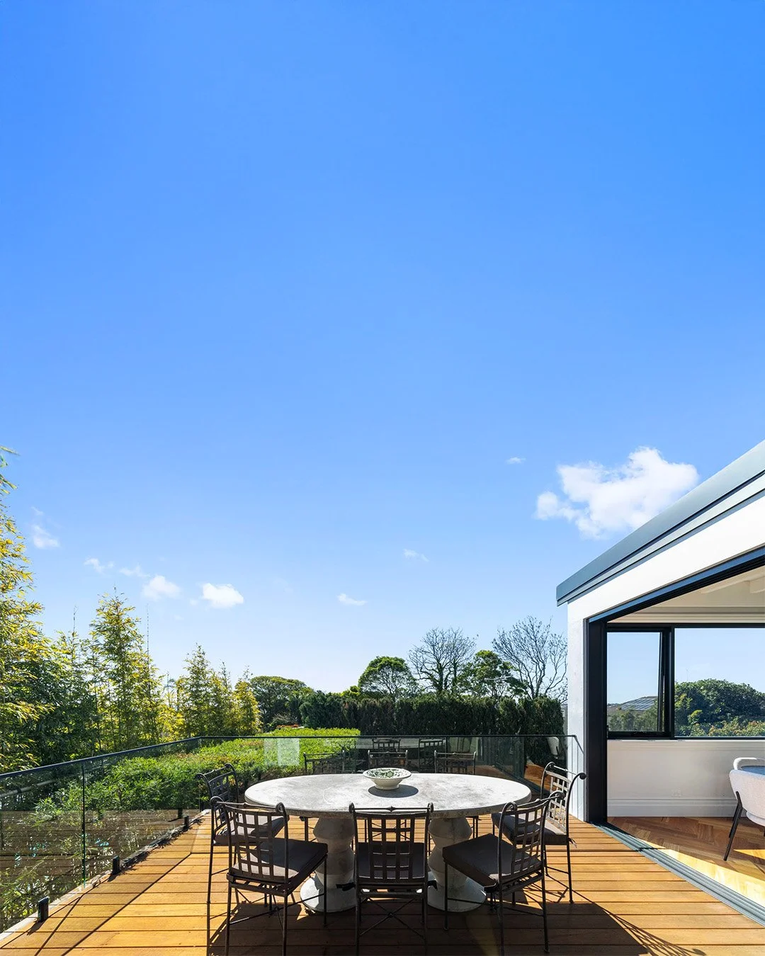 Outdoor balcony with a round dining table and six chairs, overlooking greenery and trees under a blue sky.