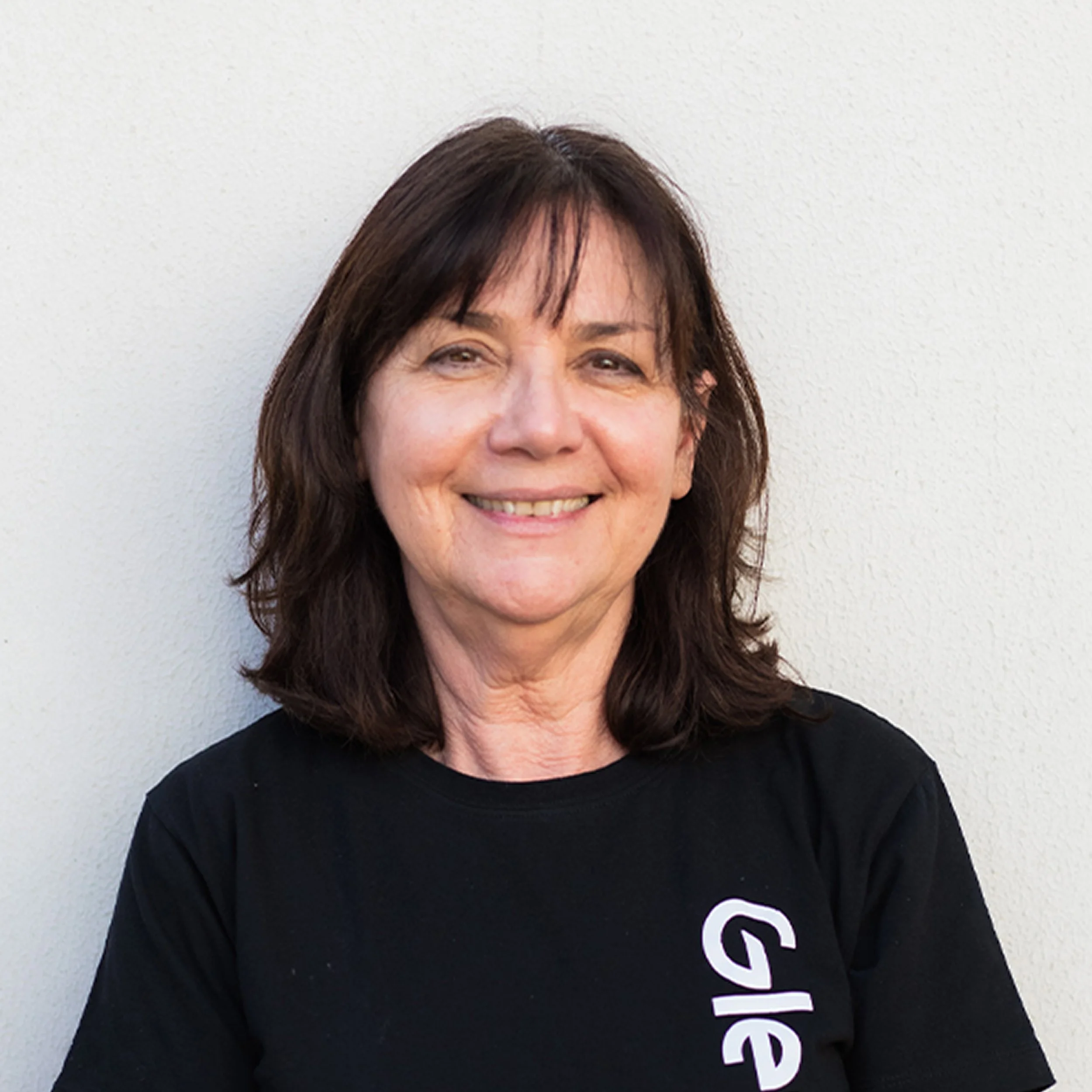 Close-up of a woman with shoulder-length brown hair, smiling, wearing a black shirt with white text, standing against a plain white wall.