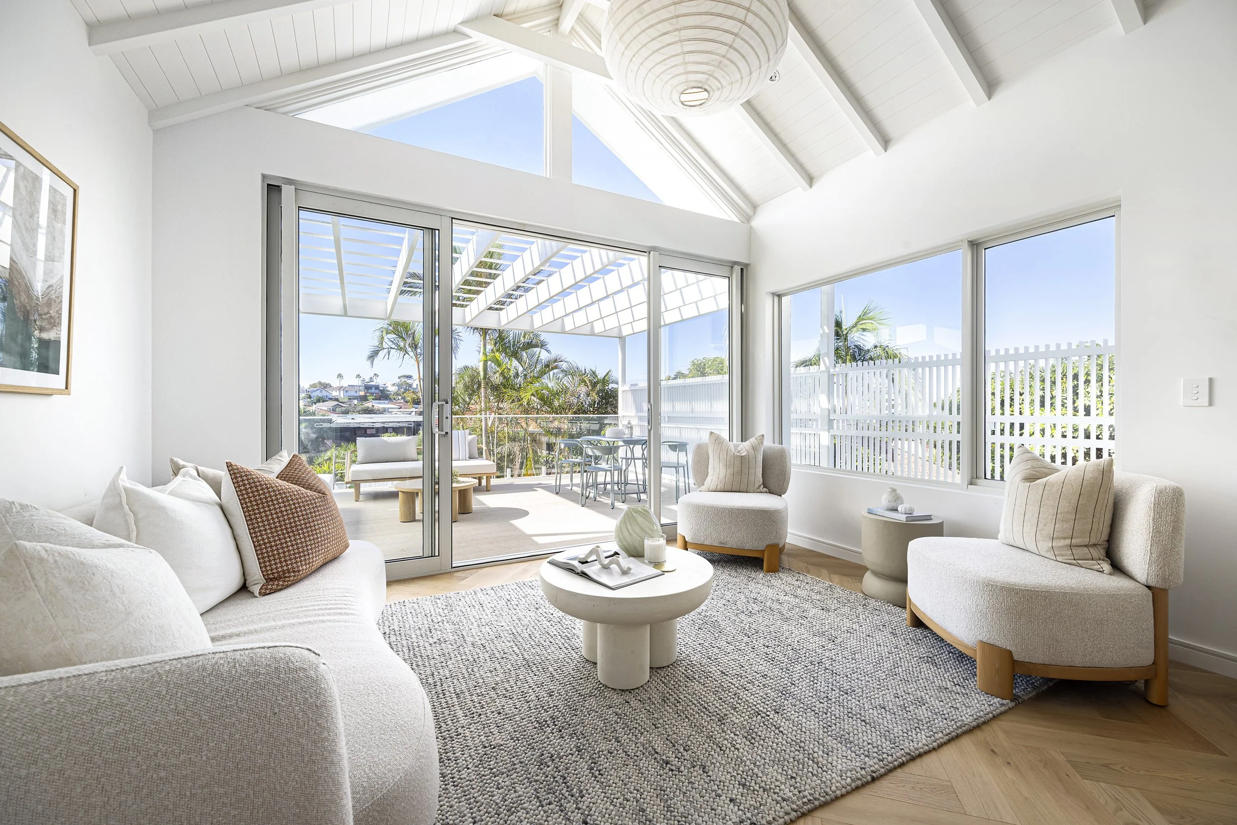 Living room with white walls and large windows showing a view of rooftops and palm trees. There is a white couch with throw pillows, a round white coffee table with decorative items, and two beige armchairs. The room has a high, vaulted ceiling with white wood beams and a hanging spherical light fixture. An open sliding glass door leads to an outdoor patio with seating and greenery.