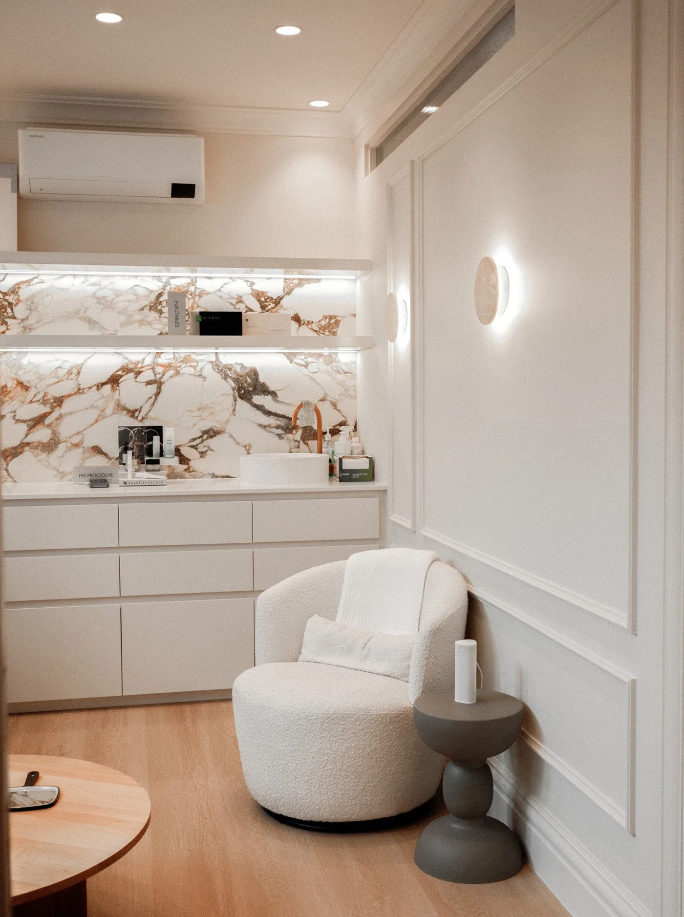 A cozy sitting area with a white plush armchair, a small round side table with an Amazon Echo, and a modern lamp with a spherical light. The background features a white kitchen counter with a marble backsplash, built-in drawers, and shelves with skincare and beauty products. The wall has white paneling and circular wall lights.