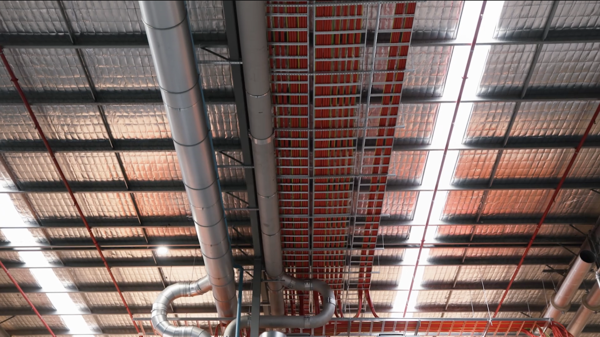 Ceiling with industrial HVAC ducts, ventilation pipes, and fire sprinkler system piping in a large commercial or industrial building.