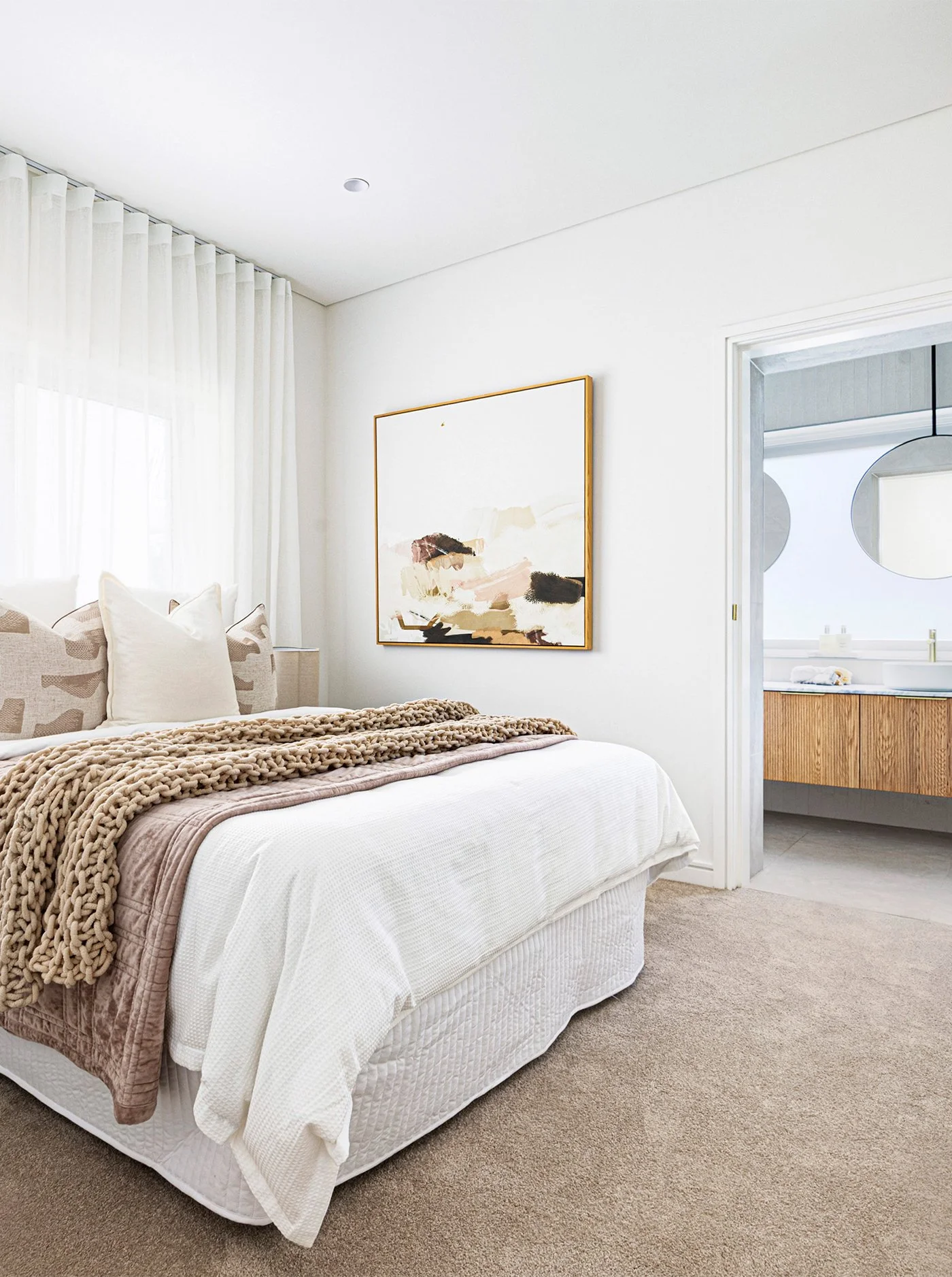 A cozy bedroom with a white bed, beige and pink throw blanket, decorative pillows, a large abstract artwork on the wall, and an adjacent bathroom with a wooden vanity and round mirror.