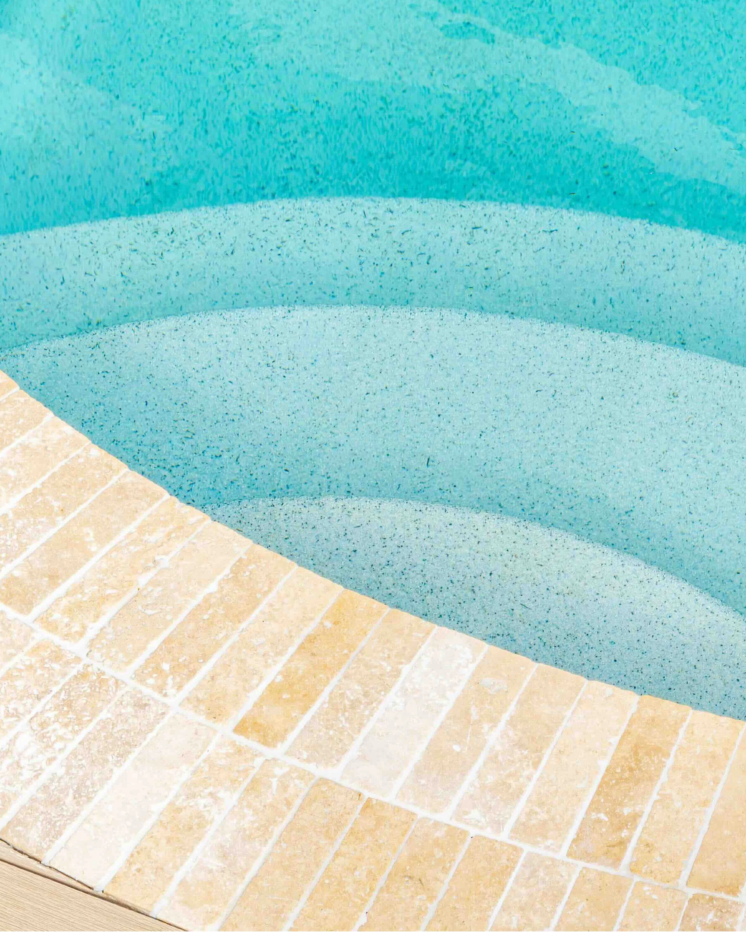 Close-up of a corner of a swimming pool with turquoise water and beige tiled edge.
