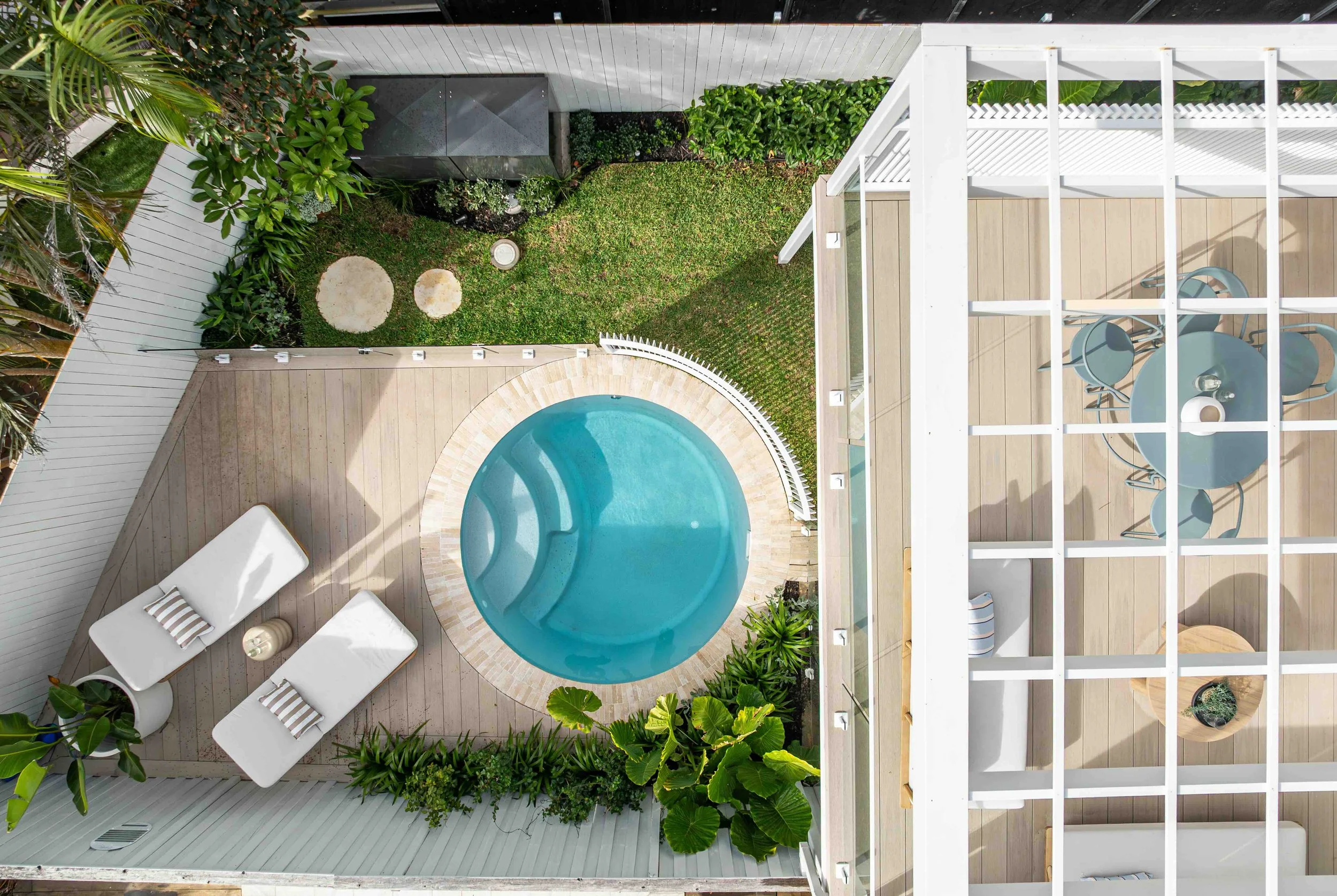 Top-down view of a backyard patio with a small swimming pool, lounge chairs, a dining table with chairs, green plants, and a white wooden fence and deck.