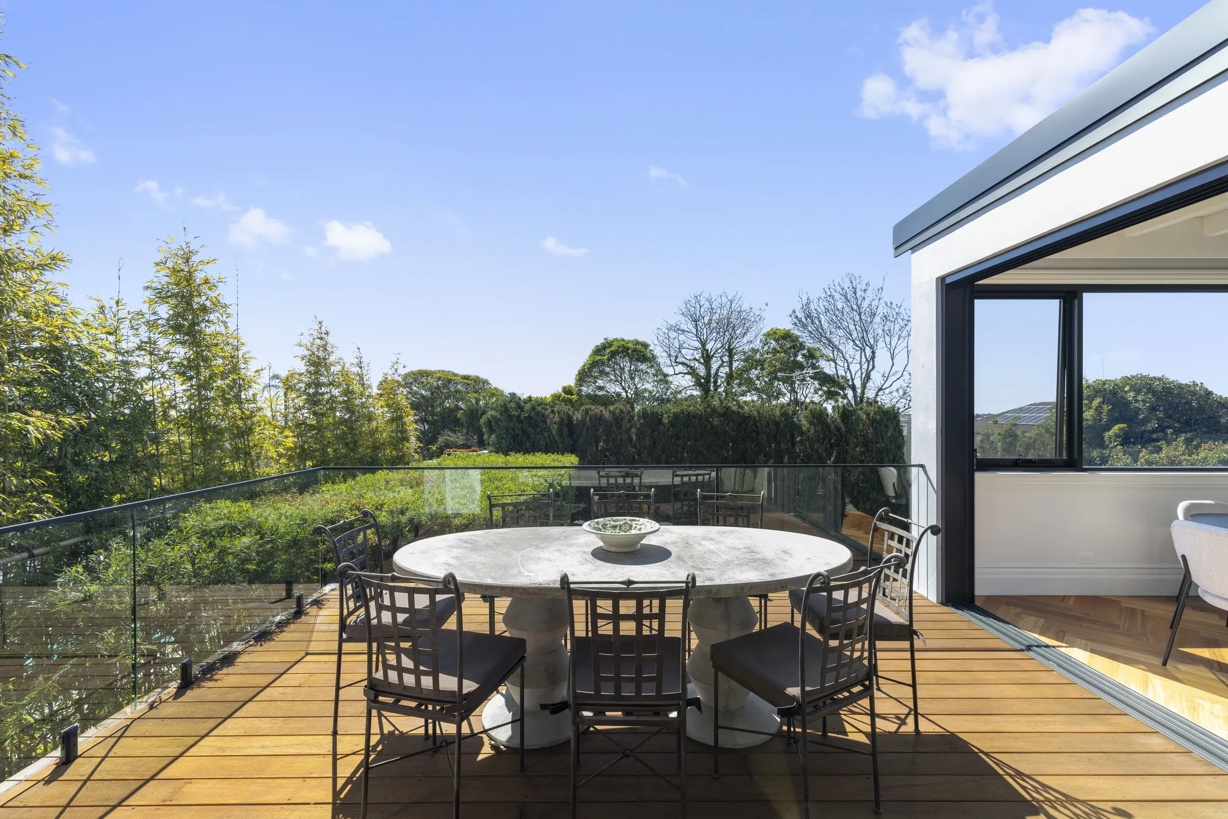 Outdoor patio area with a round table and chairs, wooden decking, a modern house with large windows, lush greenery including trees and bushes, and a clear blue sky.