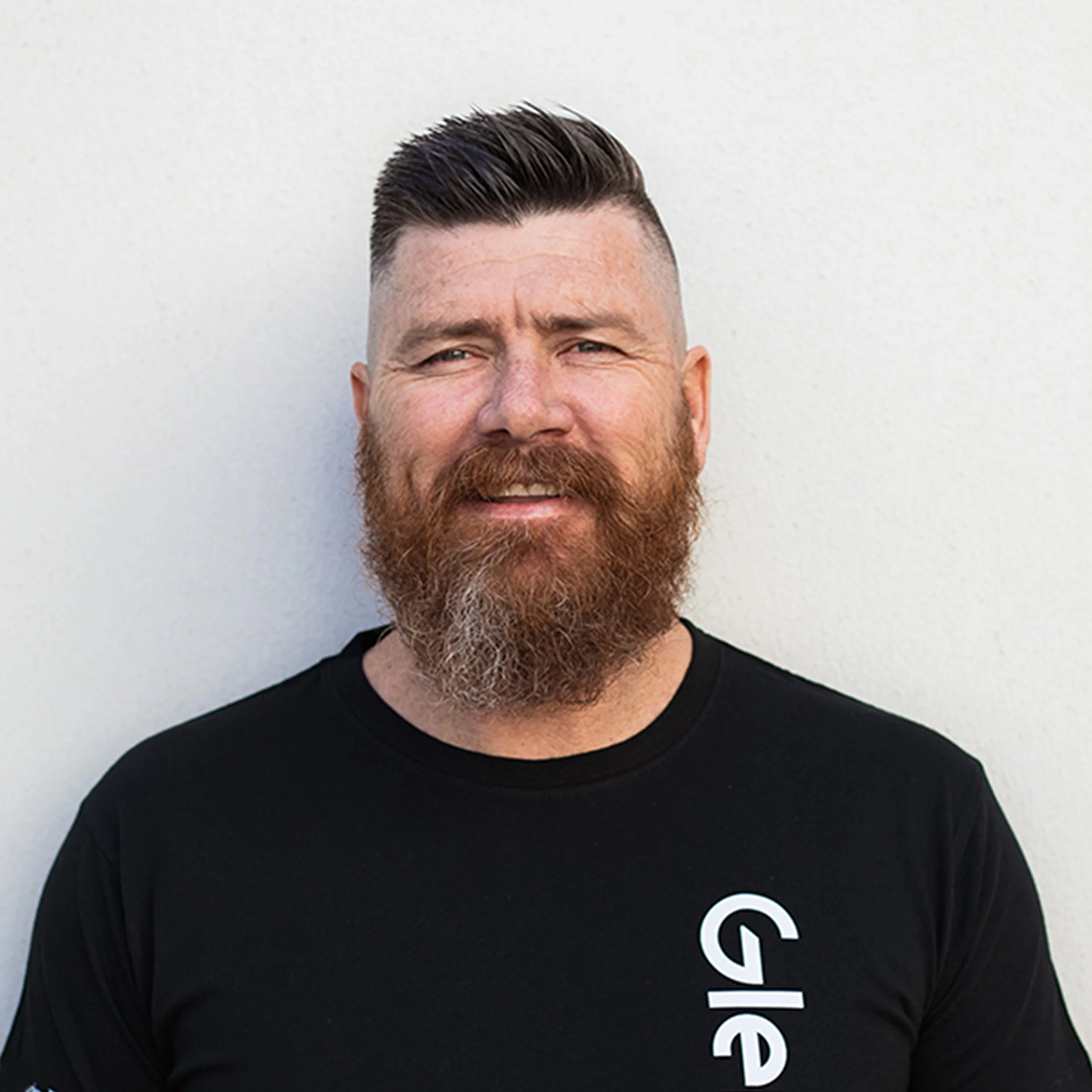 Close-up of a smiling man with dark, slicked-back hair, a beard, and wearing a black Glenco shirt, standing against a plain background.