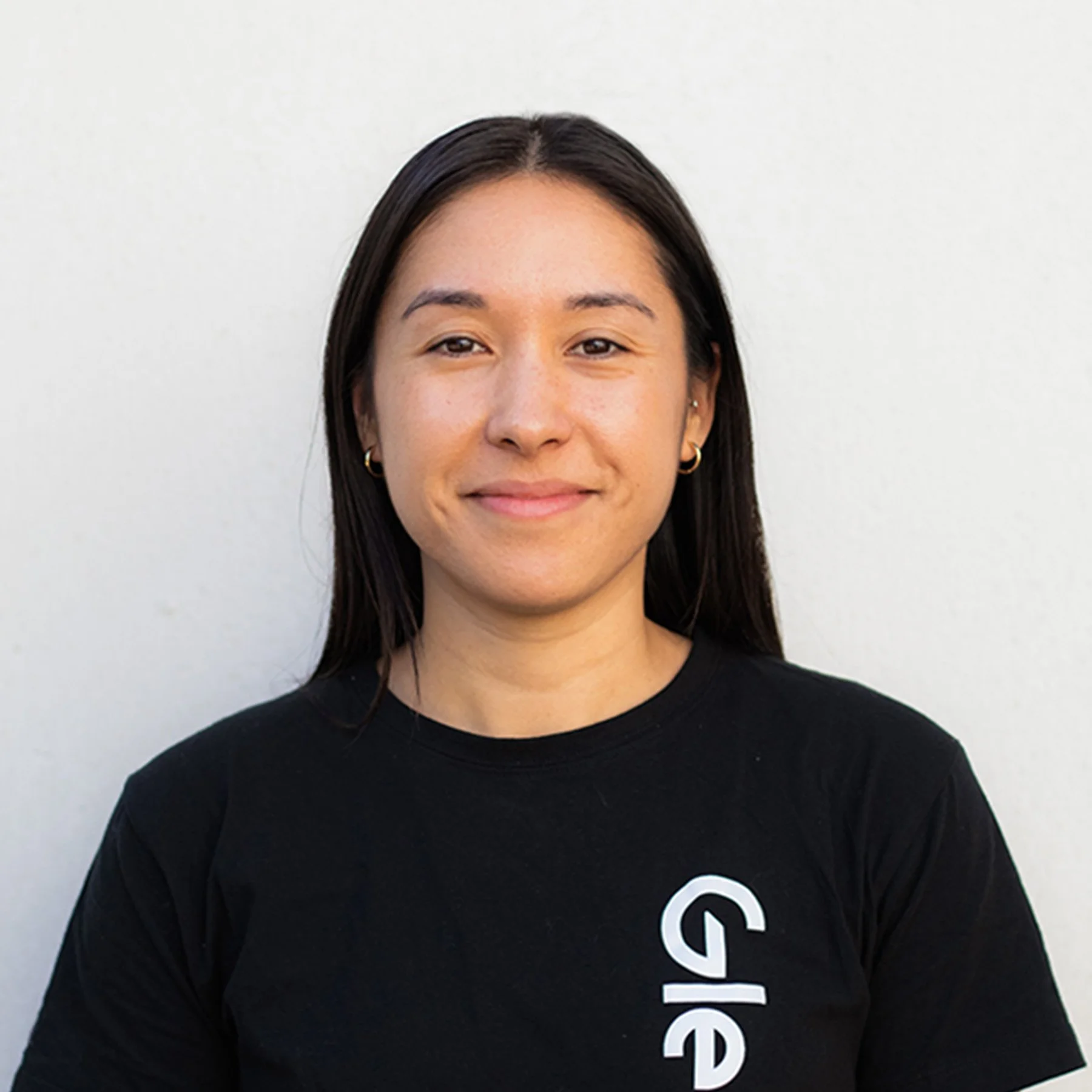 A young woman with long dark hair, smiling, wearing a black shirt with white text, standing against a plain white background.