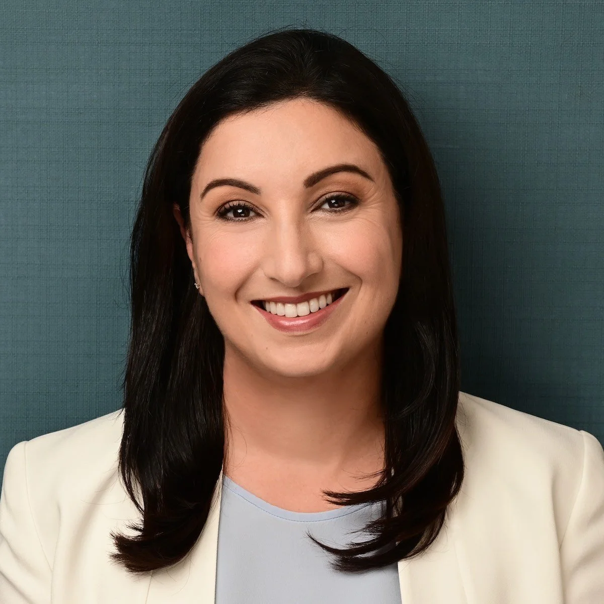 Headshot of a smiling woman with dark hair, wearing a white blazer and light-colored top, against a textured teal background.