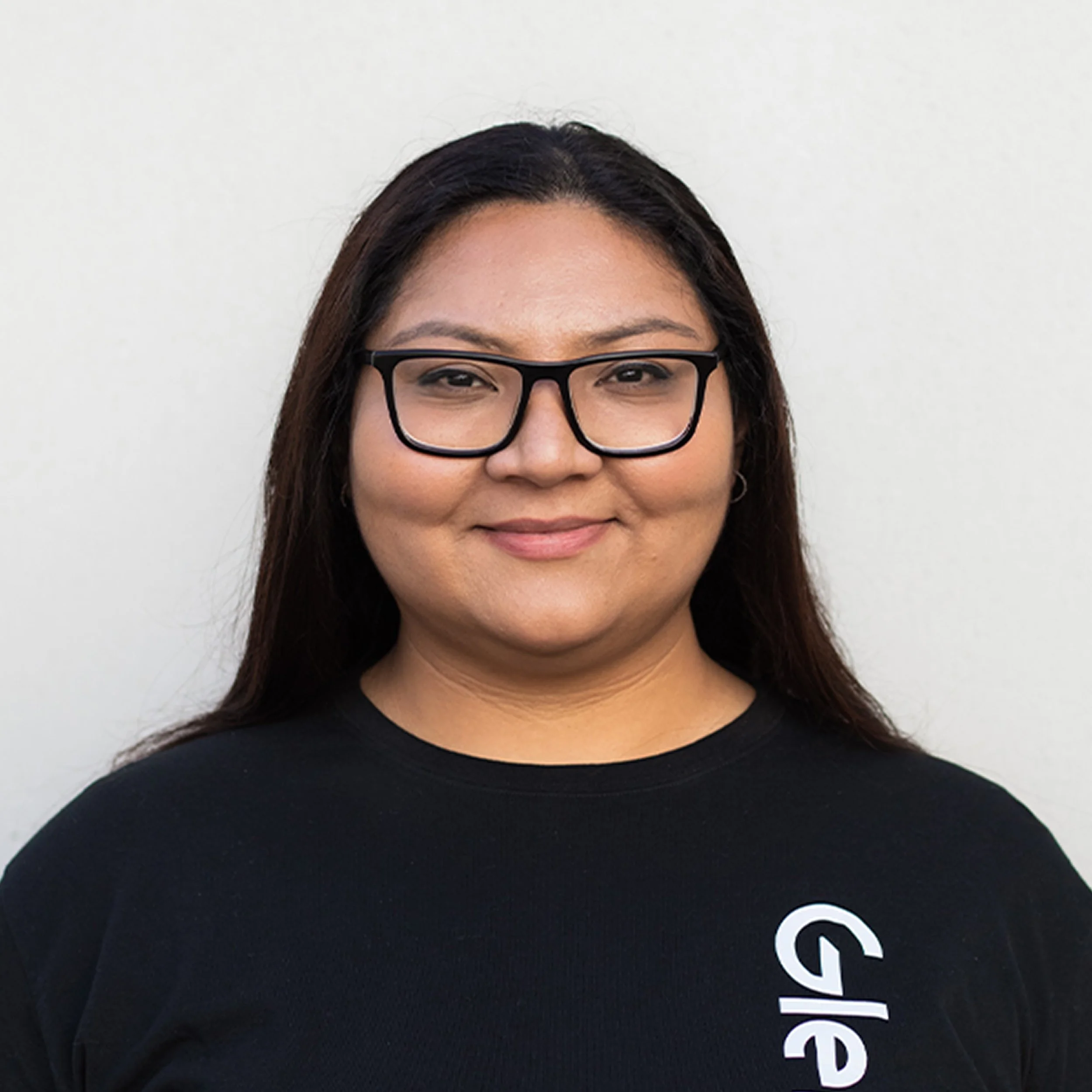 Close-up of a woman with long dark hair, wearing black glasses, smiling, against a plain white background, wearing a black shirt with white text.