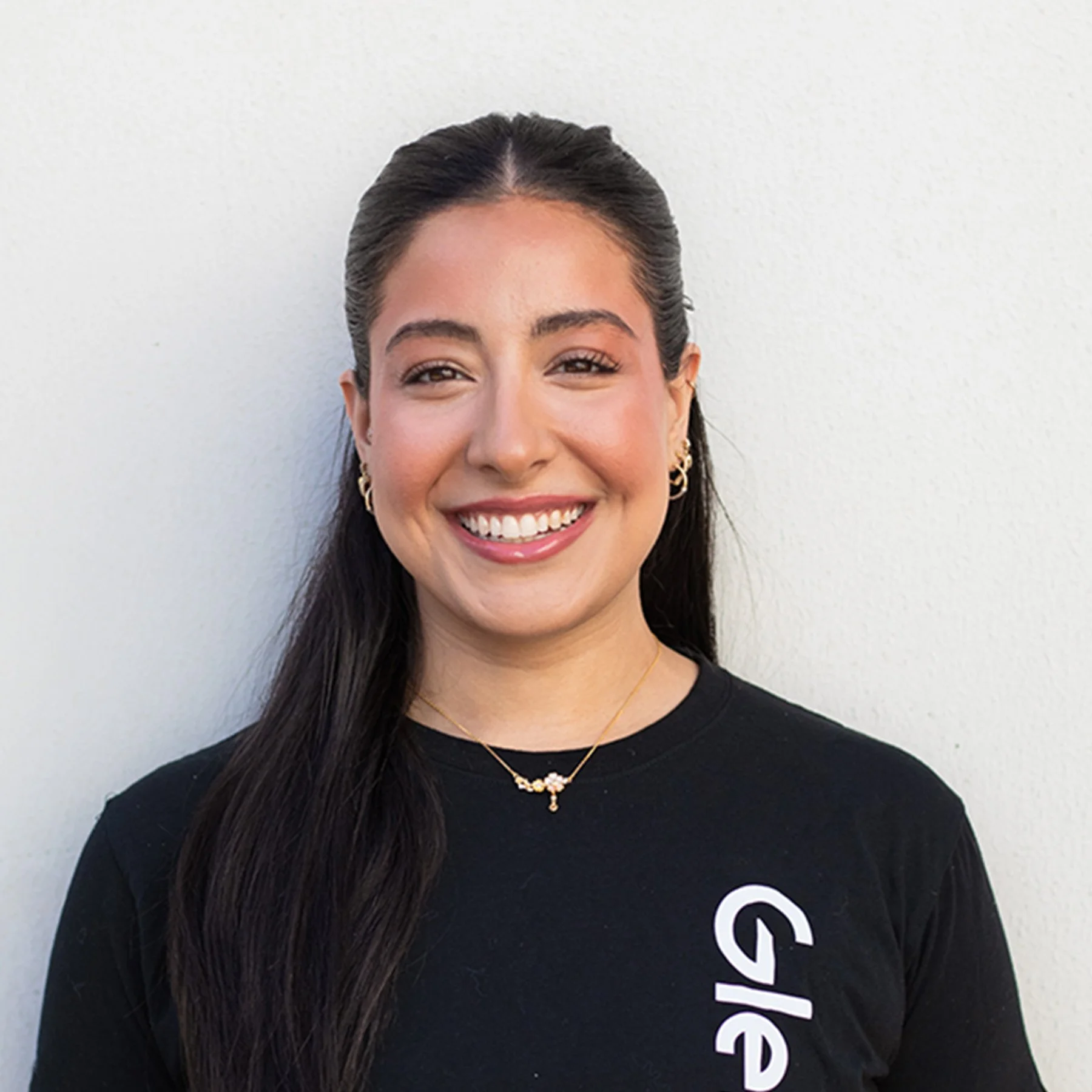 Smiling young woman with long dark hair, wearing gold earrings, smiling against a plain white wall.