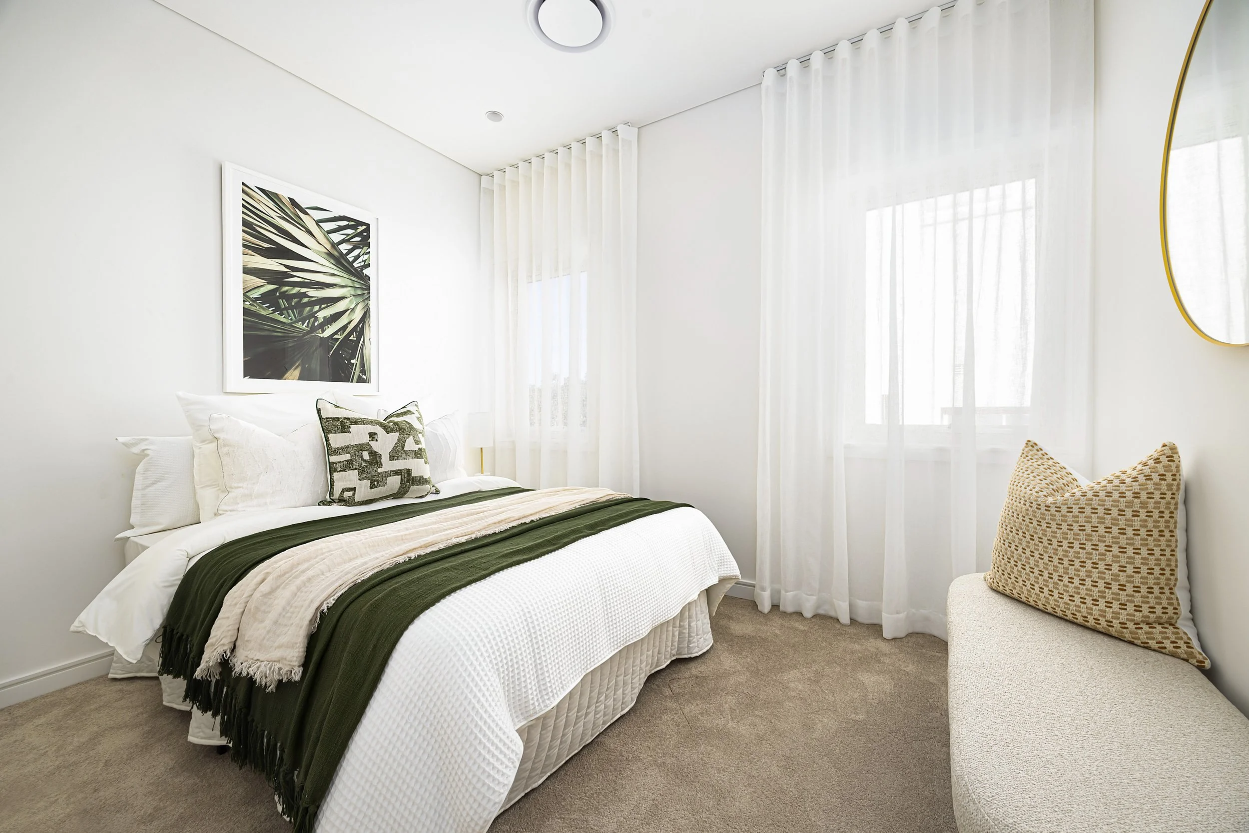 A bedroom with a white bed, green and beige bedding, black and white pillows, a framed tropical leaf print on the wall, cream curtains, and a wicker chair with a patterned pillow.