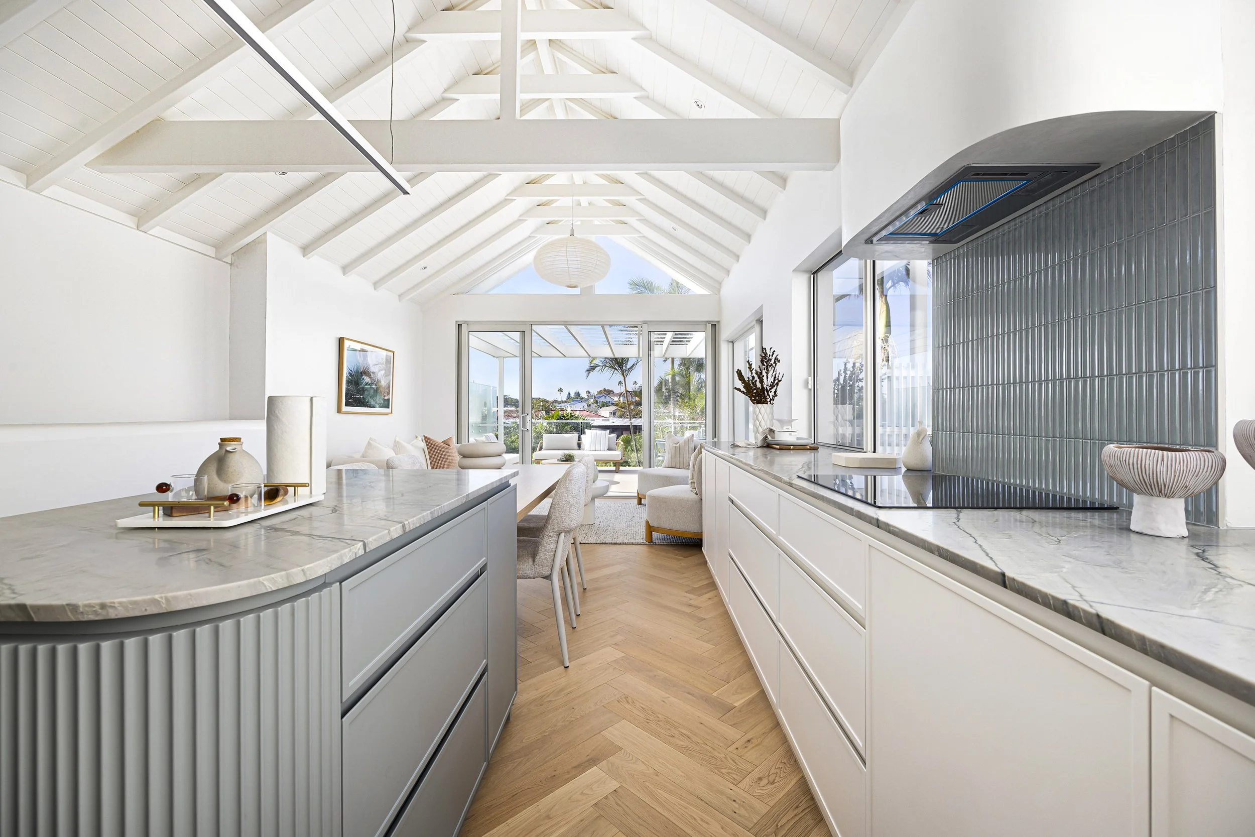 Bright, modern kitchen with white cabinets, marble countertops, and a high vaulted ceiling with white wood beams. Large windows overlooking an outdoor patio with palm trees allow ample natural light. Minimalist decor includes vases and a tray on the counter.