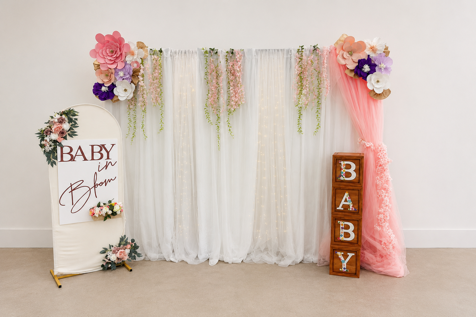 Decorative baby shower backdrop with white, pink, purple, and white flowers, pink tulle, and string lights, featuring a standing sign that reads 'Baby in Bloom' and wooden blocks spelling 'BABY' on the right.