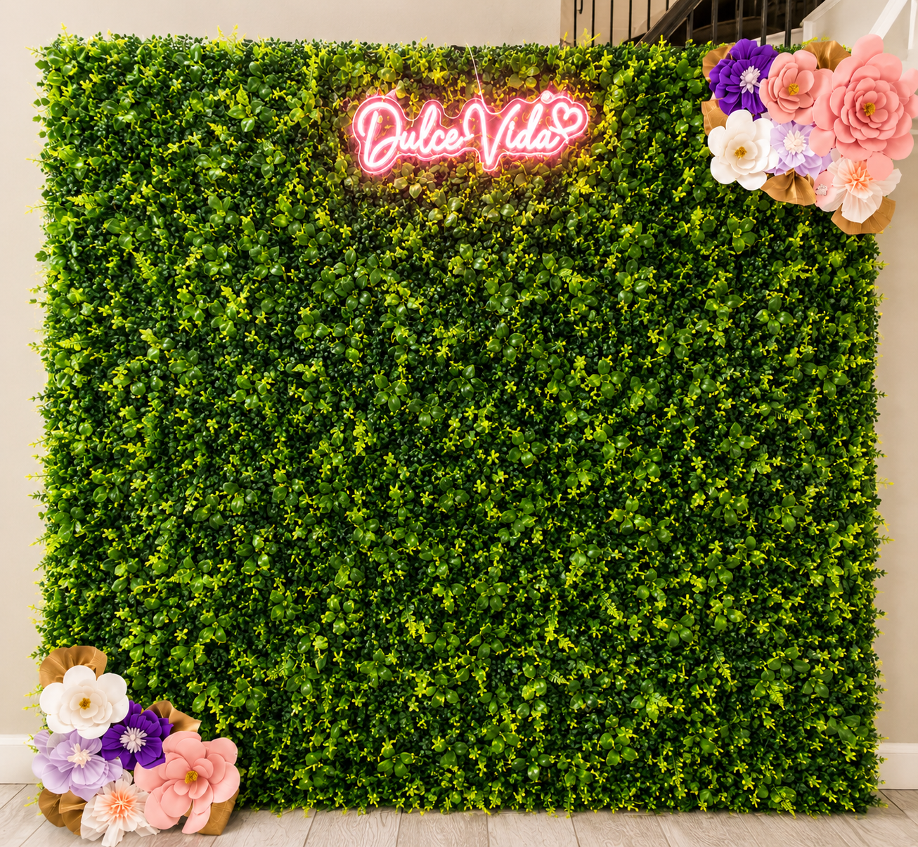 A green artificial hedge wall with a neon pink sign that reads 'Dulce Vida' and a floral arrangement with purple, pink, white, and peach-colored paper flowers on the top right and bottom left corners.