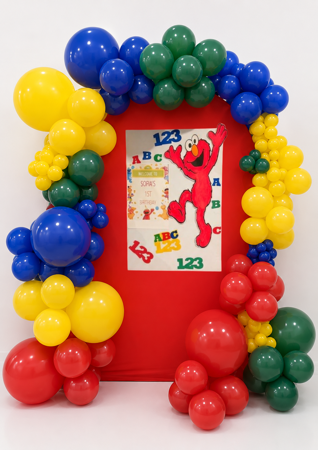 Colorful balloon arch in blue, yellow, red, and green surrounds a Sesame Street birthday poster for Sofia's 1st birthday, with a picture of Elmo and alphabet and number decorations.