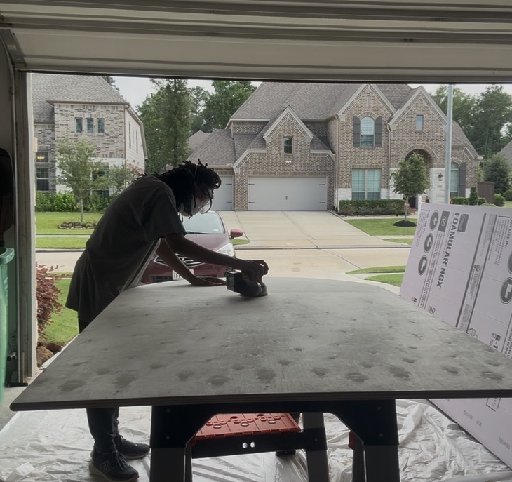 Person using a handheld power tool on a large table in a garage, with a residential neighborhood visible outside.