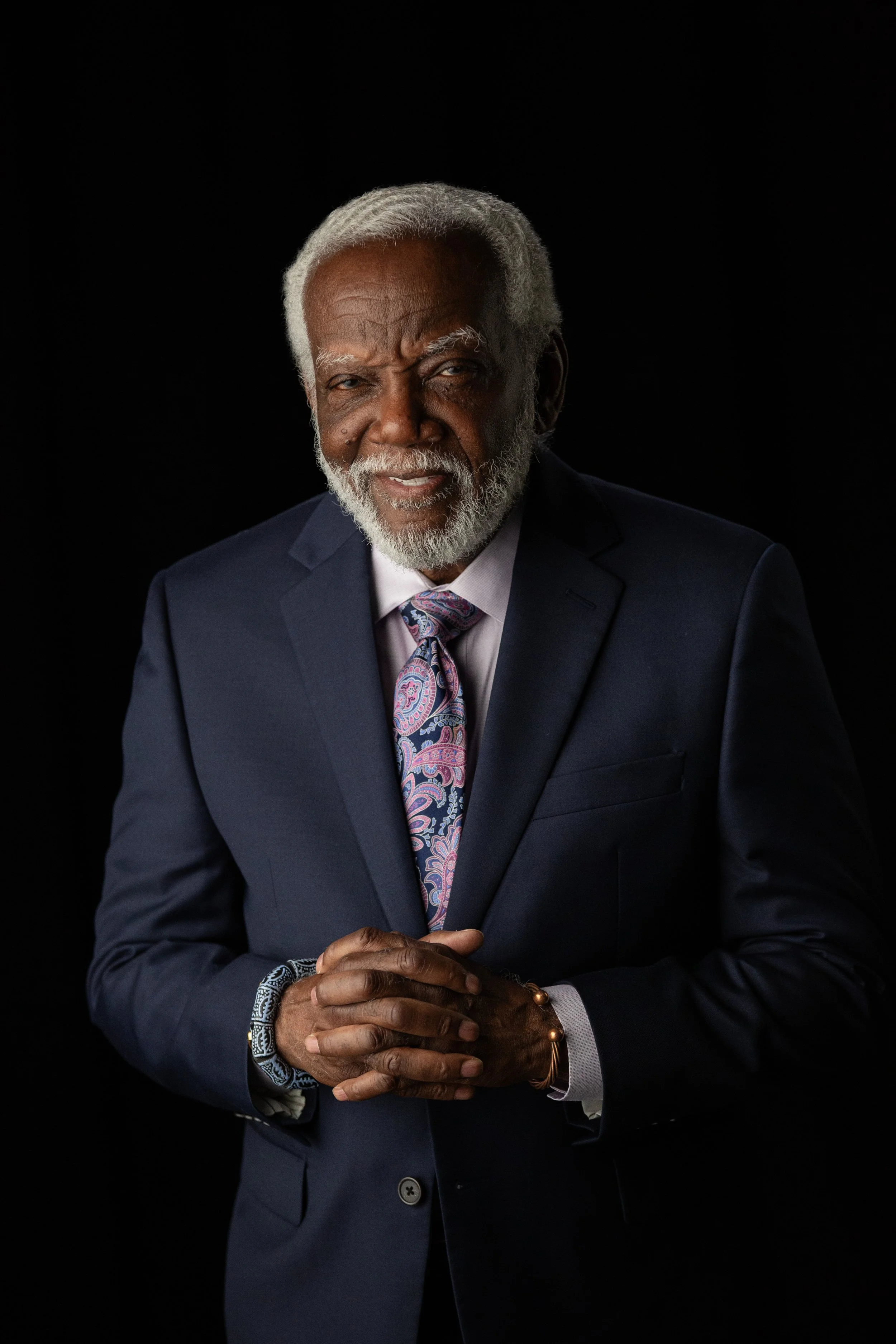 Portrait of an elderly African American man with gray hair and a beard, wearing a navy blue suit, a paisley tie, and jewelry, posing against a black background.