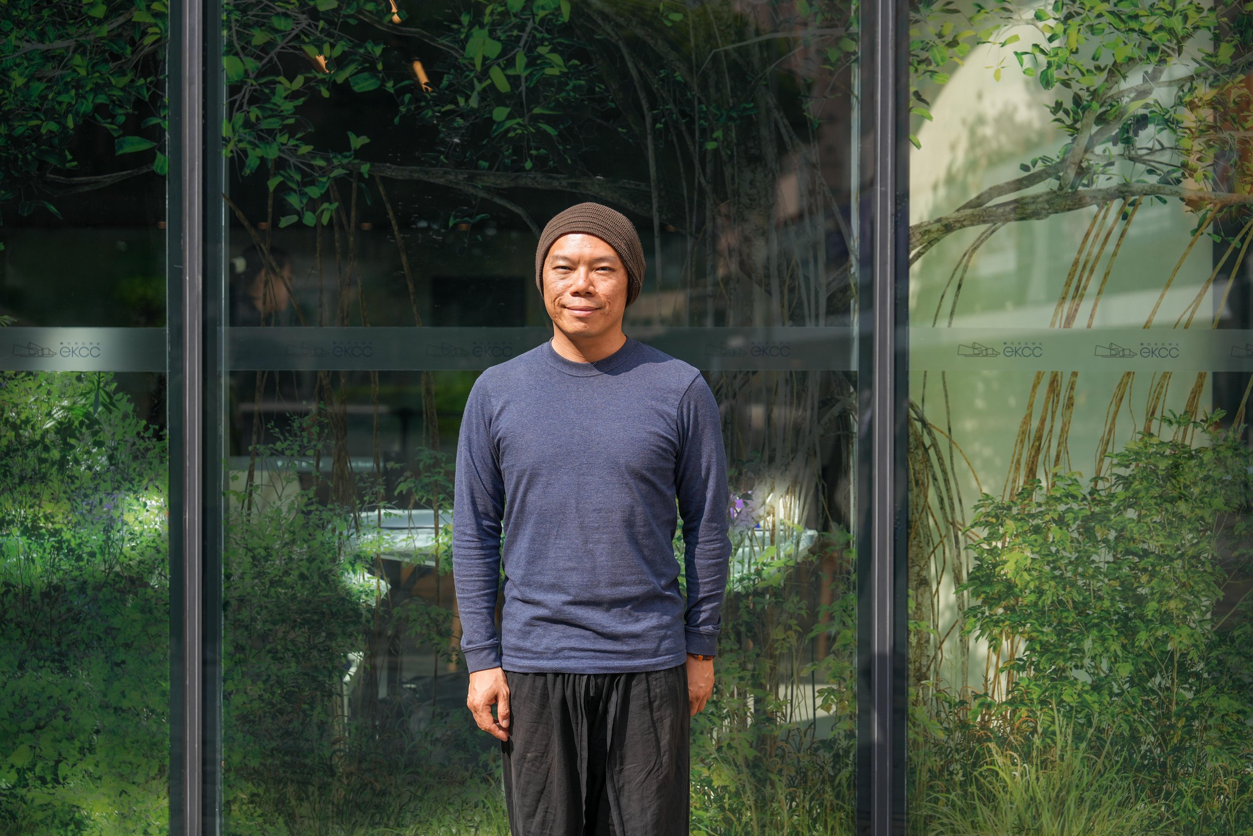 A man wearing a brown beanie and a long-sleeved blue shirt stands outdoors in front of a glass barrier with green plants behind it.