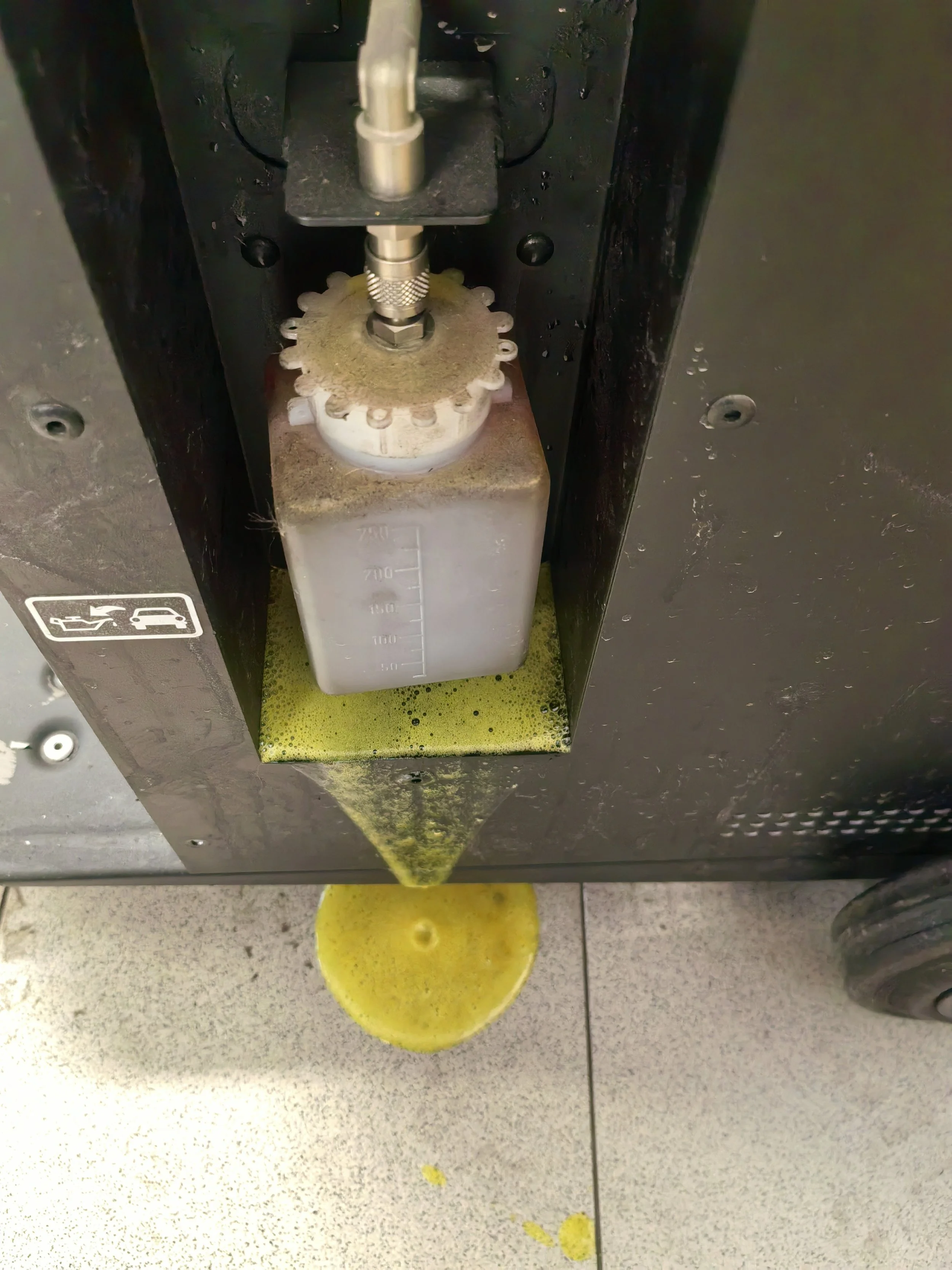Máquina de llenado de líquidos con un recipiente transparente y medidor, vertiendo líquido en un recipiente, en un área de laboratorio o industrial.