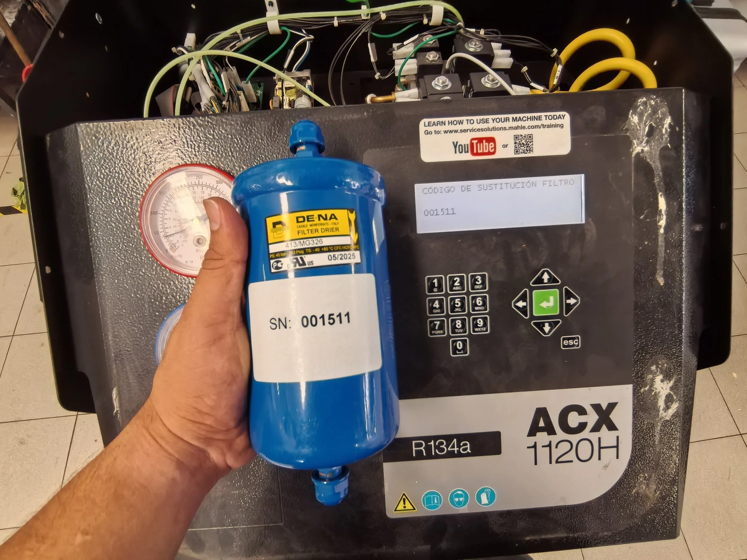 Persona sosteniendo un filtro de refrigerante azul frente a una caja de aire acondicionado con cables y componentes internos visibles.