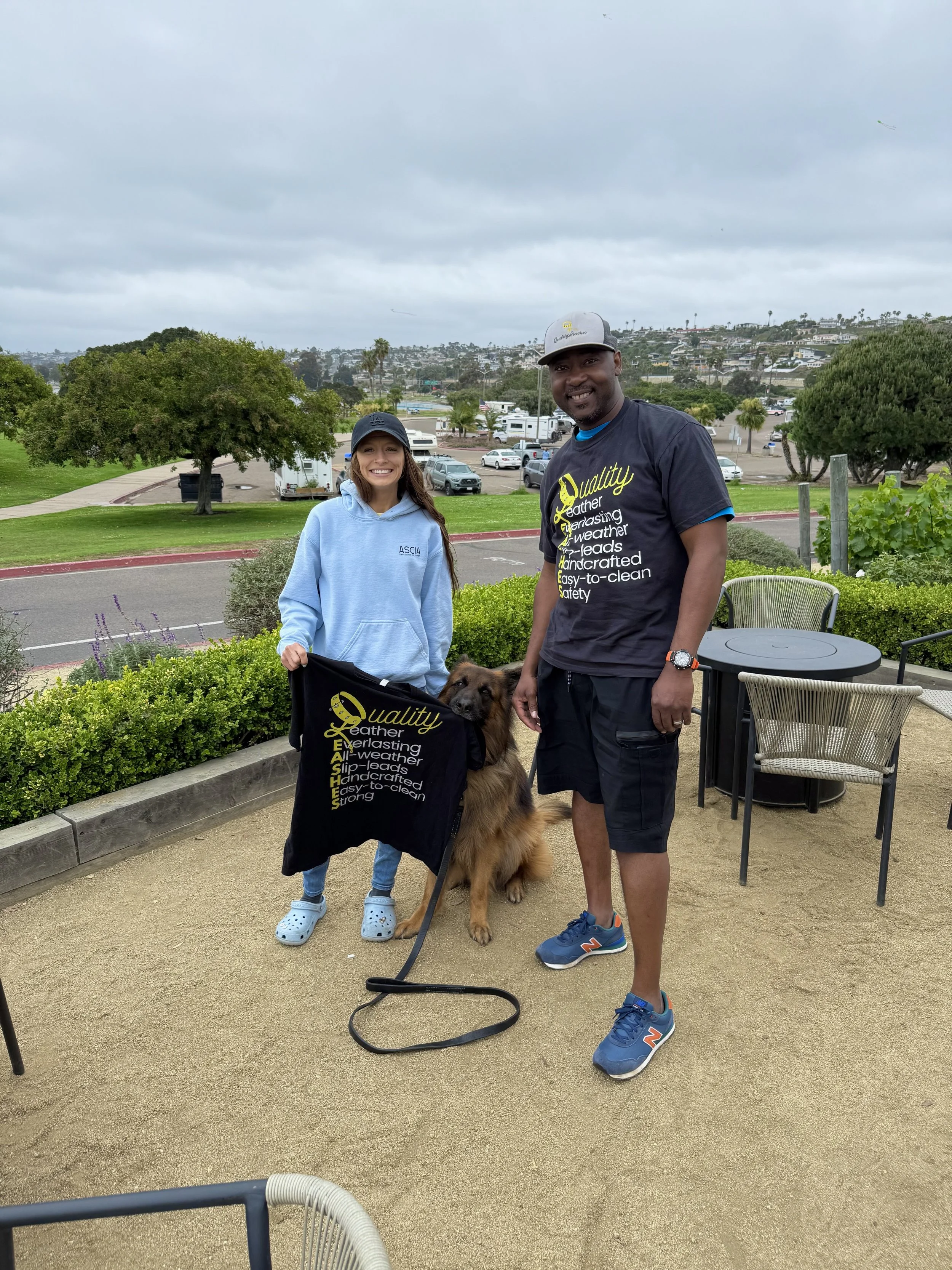 Two smiling people with a large German Shepherd dog standing between them outdoors. The woman is wearing a light blue hoodie and black cap, and the man is wearing a black t-shirt, gray cap, and blue sneakers. Both are holding a black Chelsea football club flag. The background includes trees, parked cars, RVs, and a hill under a cloudy sky.