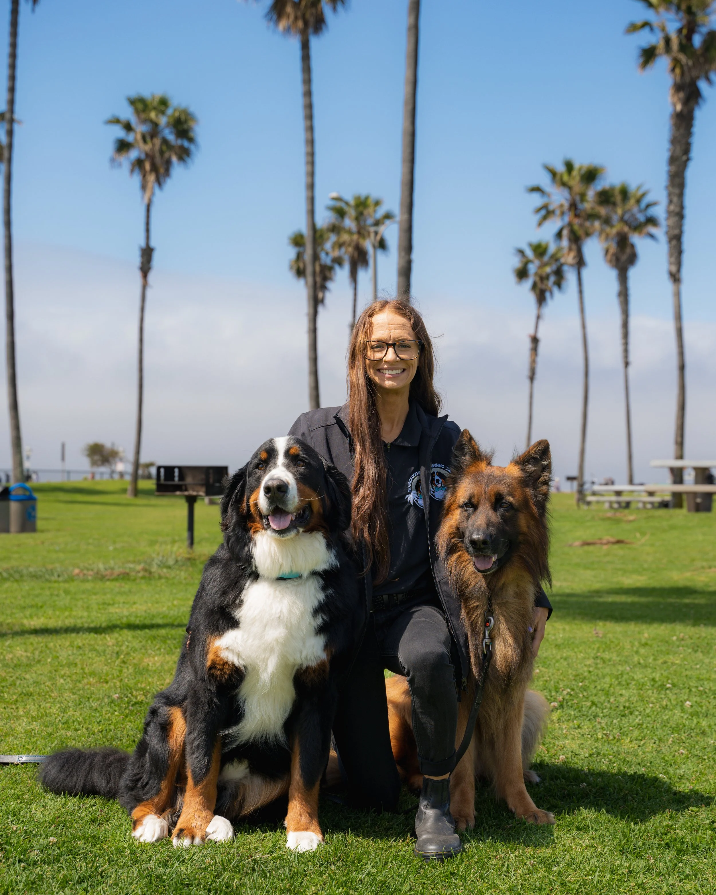 A woman with glasses smiling, kneeling on grass with three dogs in a park with palm trees and a blue sky in the background.