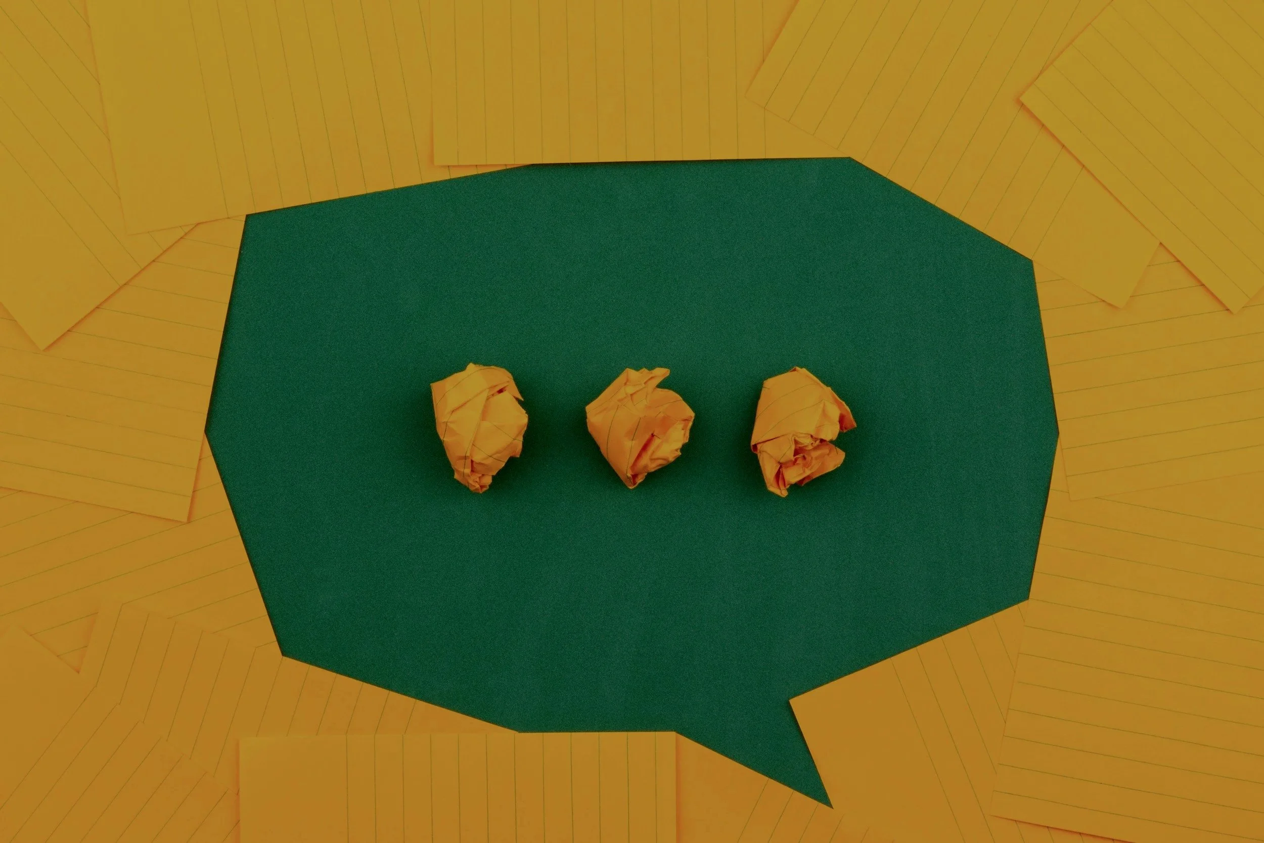 Yellow sheets of paper arranged around a green speech bubble cut from a piece of paper with three crumpled yellow balls on top.