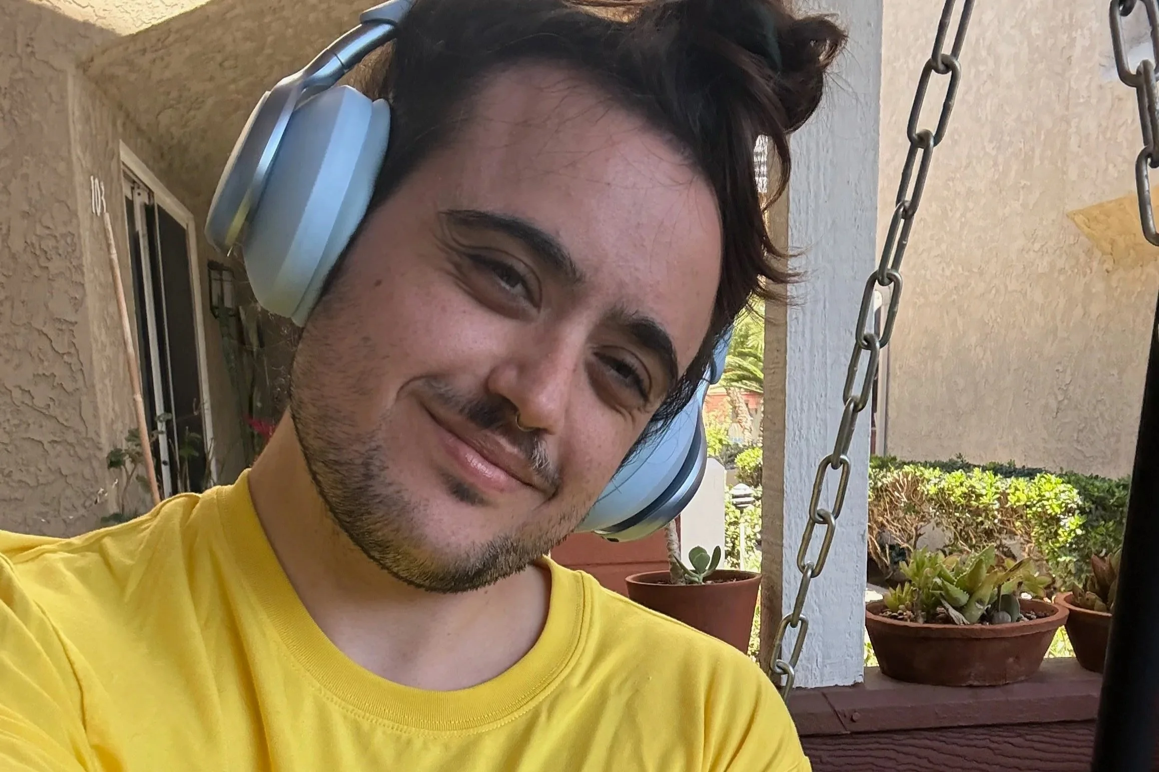 A young man with dark hair, facial hair, and glasses, smiling while wearing large headphones and a yellow shirt, sitting on a porch with plants, trees, and a building in the background.