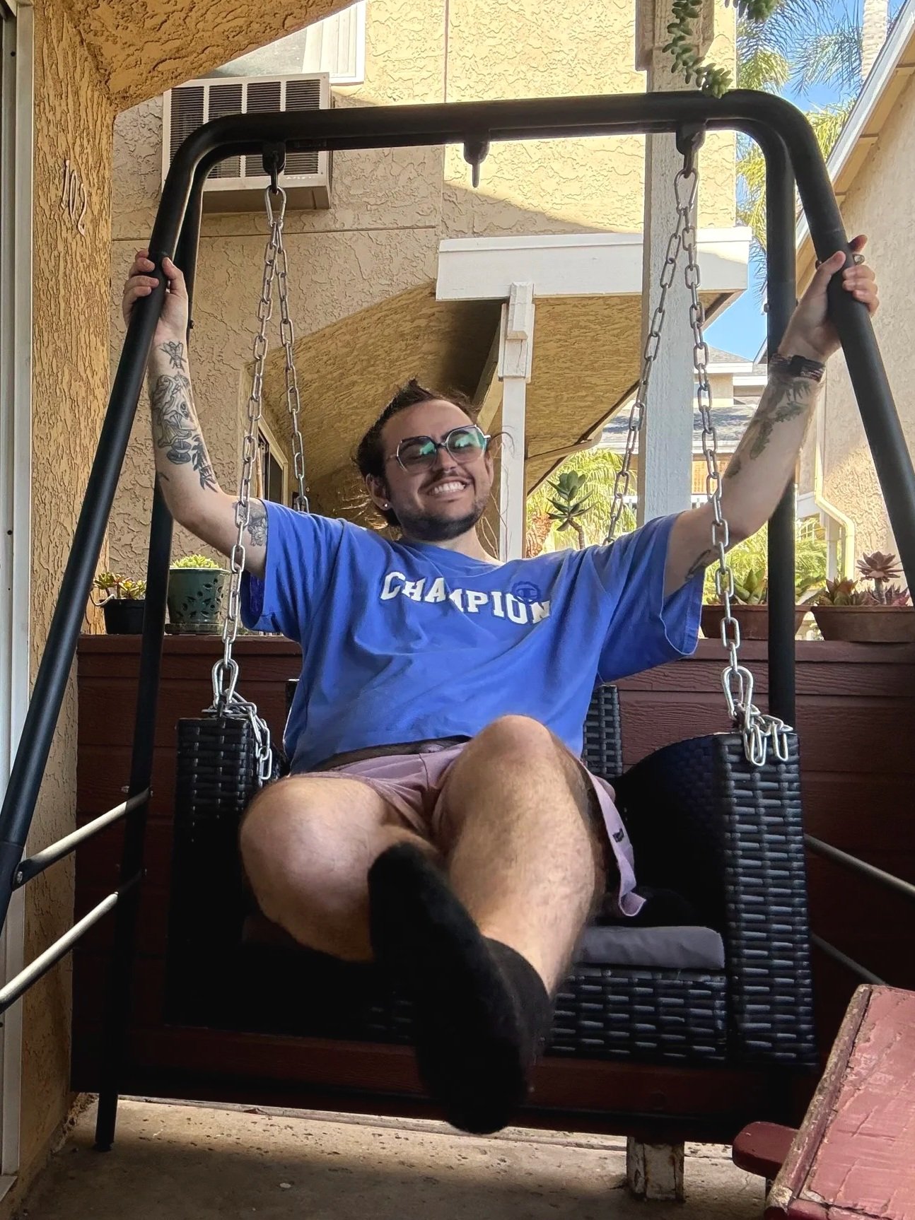 A smiling person with glasses and tattoos on their arms sitting on a swing with a happy expression, outdoors on a patio with plants and a house in the background.