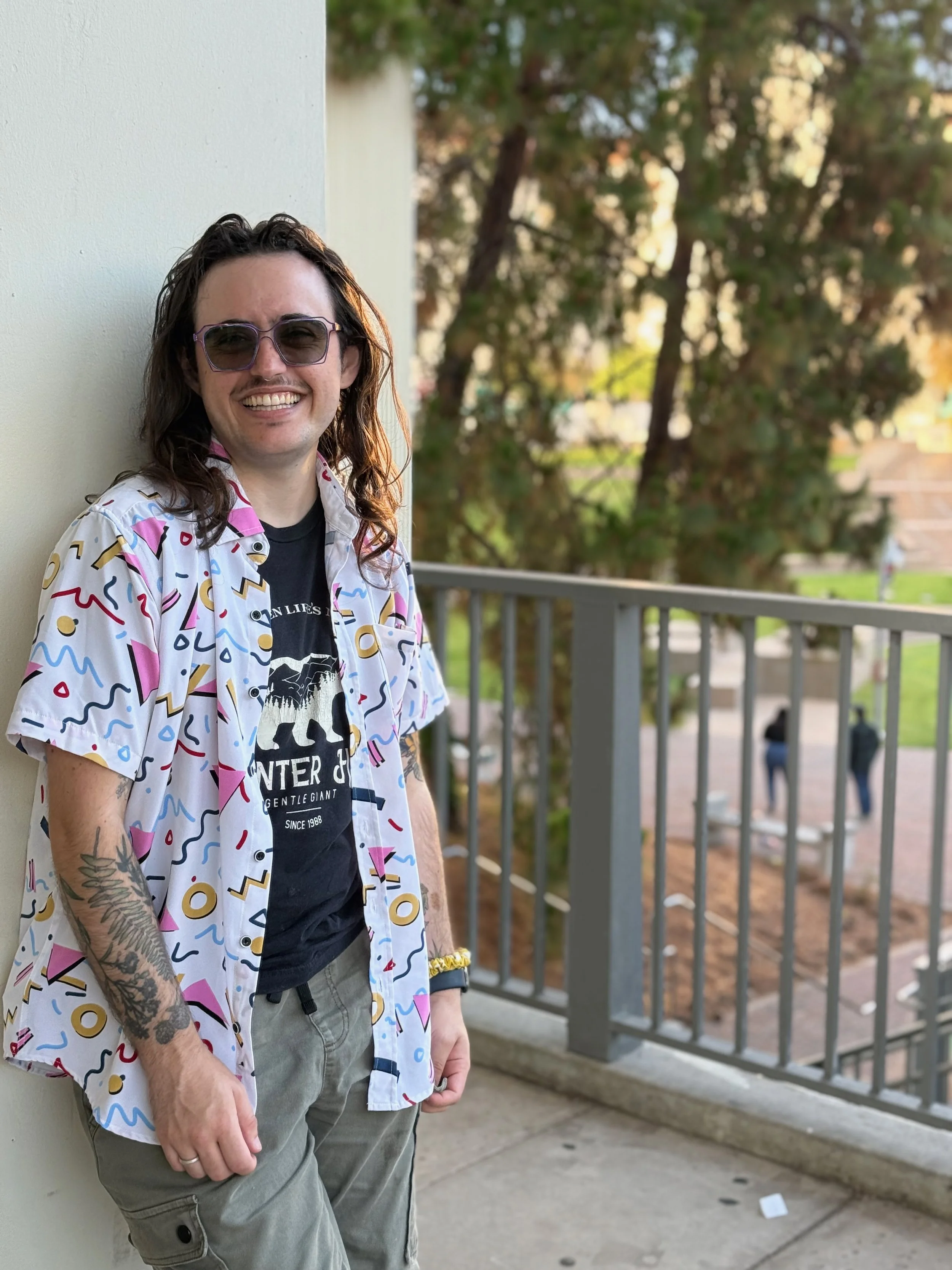 A smiling young man with long hair, wearing sunglasses, a colorful patterned shirt, and gray cargo pants, is standing next to a white wall outdoors with green trees and a metal railing in the background.