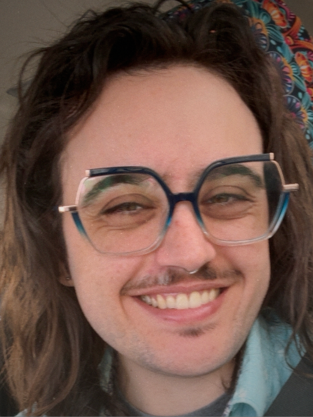 Close-up of a smiling person with long brown hair, wearing large blue glasses, and a light blue shirt.