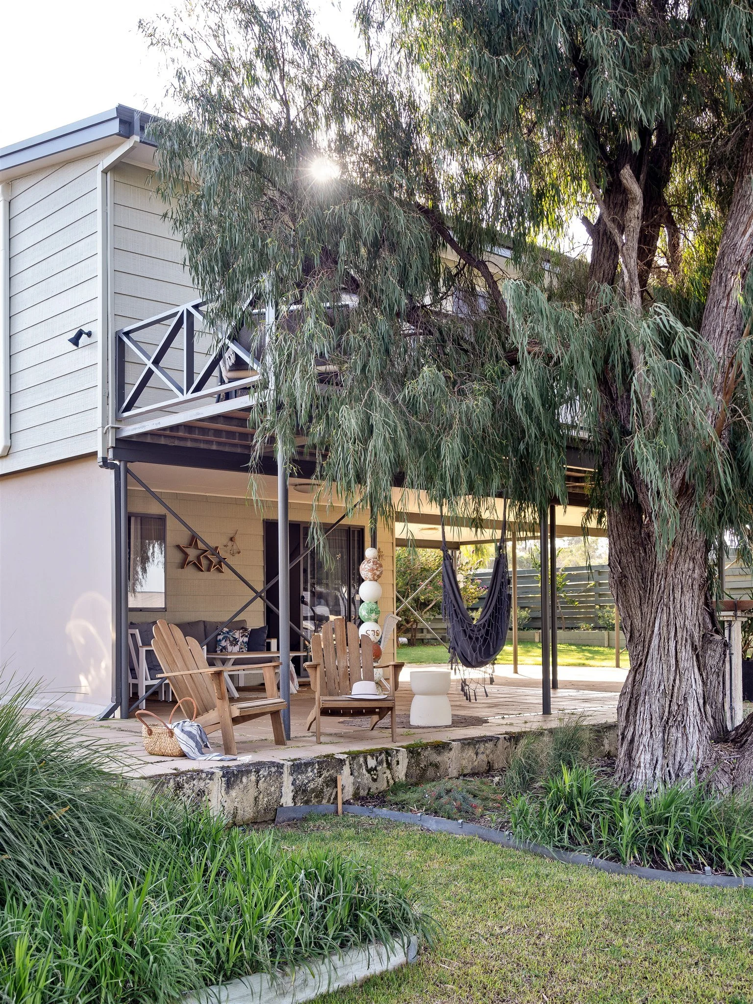 A backyard patio area with wooden chairs, a hammock, and potted plants under a large tree. The house has a balcony, and sunlight filters through the tree leaves.